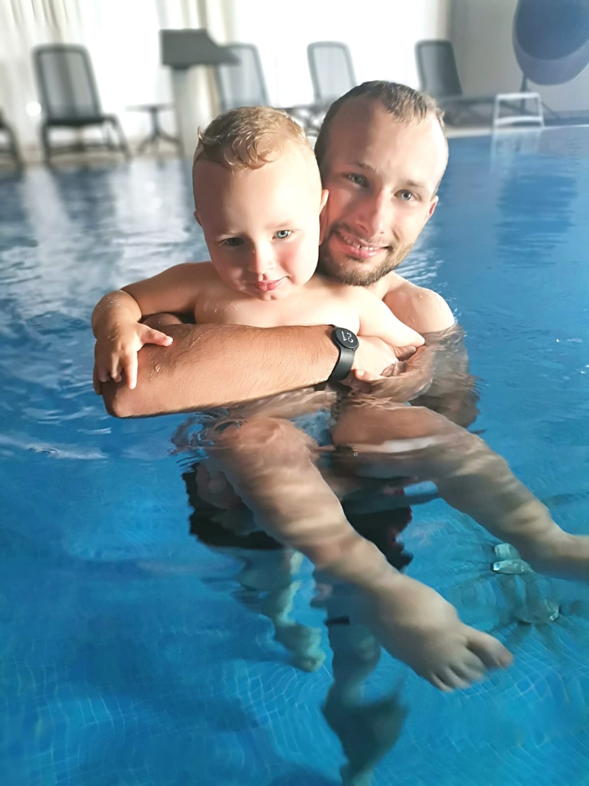 Uśmiechnięty mężczyzna z brodą trzymający małego chłopca w basenie z błękitną wodą i białymi kafelkami w tle, widoczny zegarek na ręce mężczyzny.