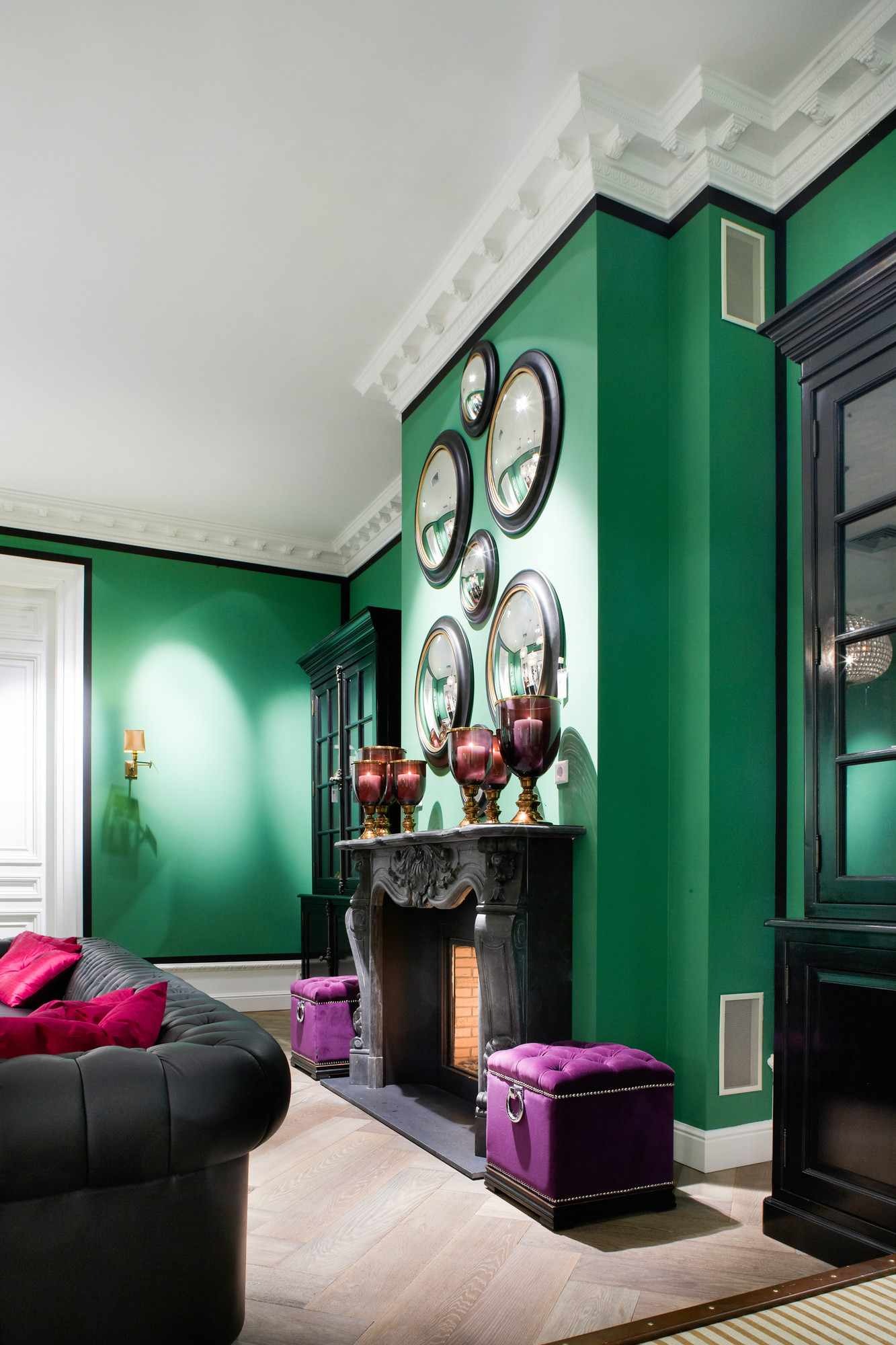 Szmaragdowozielony salon z czarnym kominkiem ozdobionym szklanymi świecznikami, kolekcją okrągłych luster na ścianie, czarną sofą z różowymi poduszkami i fioletowymi pufami.