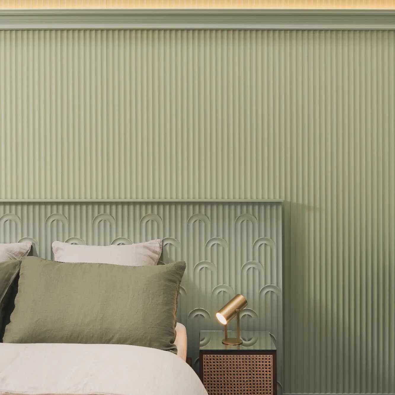 Sypialnia w odcieniach zieleni z panelem ściennym z pionowymi żłobieniami i zagłówkiem z motywem łuków, oświetlona złotą lampką nocną na plecionym stoliku.