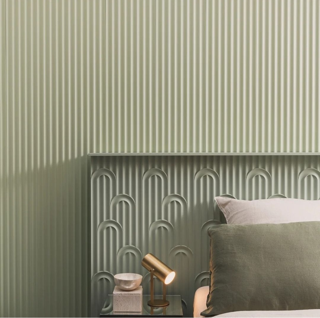 Pudrowo zielona sypialnia z panelem ściennym z pionowymi żłobieniami i dekoracyjną listwą z motywem łuków, oświetlona złotą lampką nocną na szklanym stoliku.