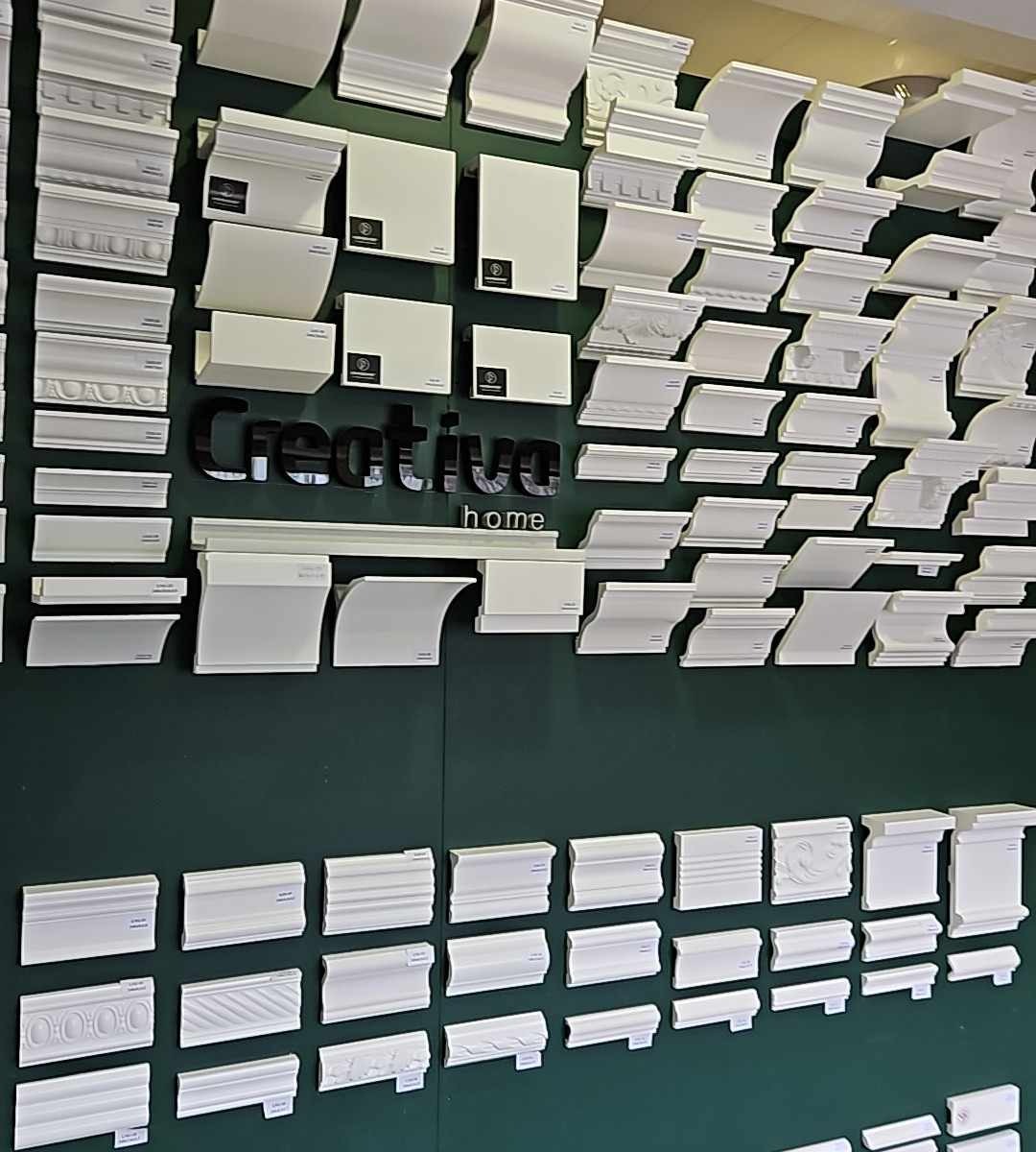 Ekspozycja różnorodnych listew przypodłogowych i sufitowych w kolorze białym, przymocowanych do ciemnozielonej ściany, z logo firmy 'Creative home' w górnej części.