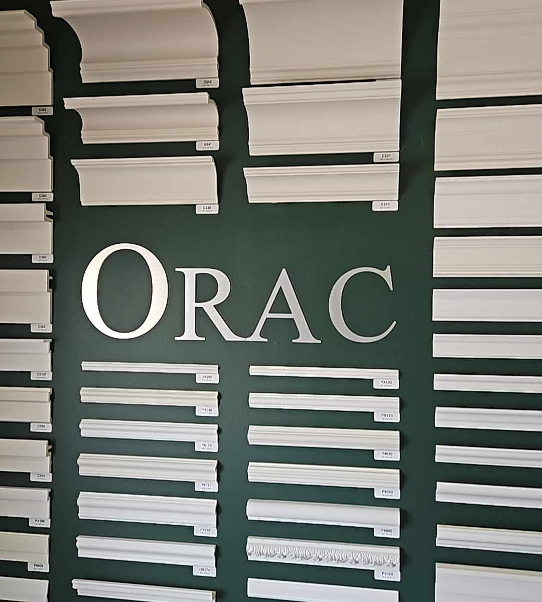 Ekspozycja różnych profili sztukateryjnych marki ORAC na ciemnozielonej ścianie, z widocznymi numerami katalogowymi poszczególnych modeli.