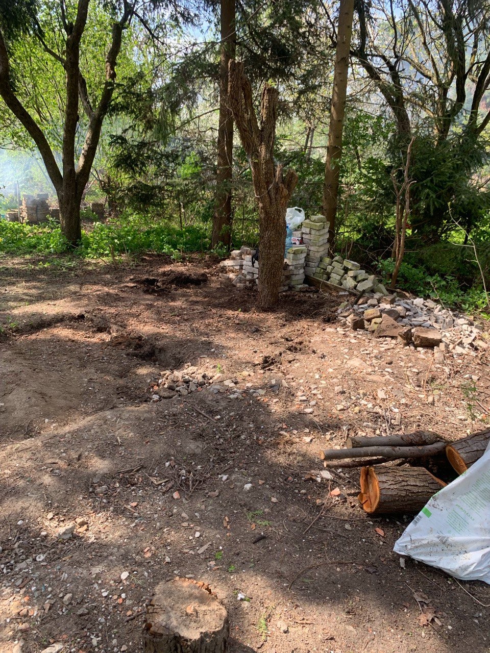 Przygotowany teren pod przyszły ogród, widoczne pniaki, powalone gałęzie, sterta kamieni i worki z ziemią, w tle drzewa i dym z ogniska.