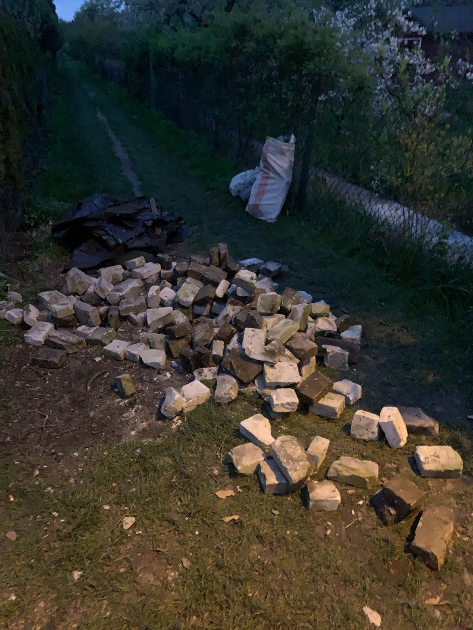 Stos starych, szarych i brązowych cegieł na ziemi w ogrodzie, obok czarnych płyt i worków z odpadami, w tle ścieżka i zieleń.