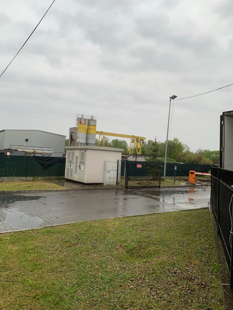 Widok na teren przemysłowy w pochmurny dzień po deszczu, z widoczną trawą, budynkiem z silosami i bramą wjazdową.