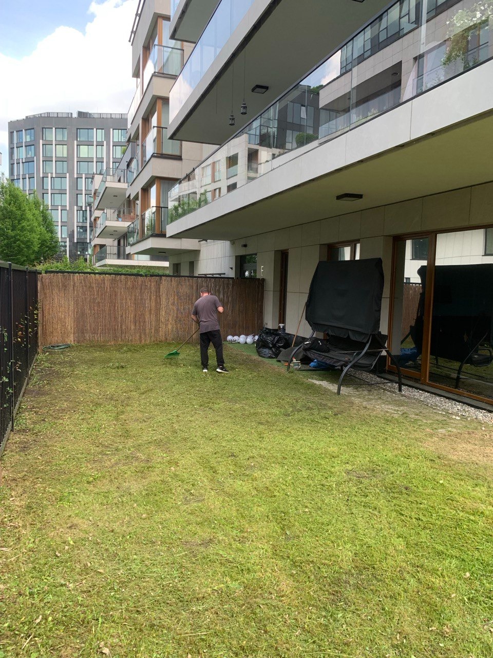 Mężczyzna grabi skoszoną trawę na trawniku przed nowoczesnym budynkiem mieszkalnym, widoczne ślady po koszeniu, ogrodzenie z wikliny i sprzęt ogrodowy w tle.