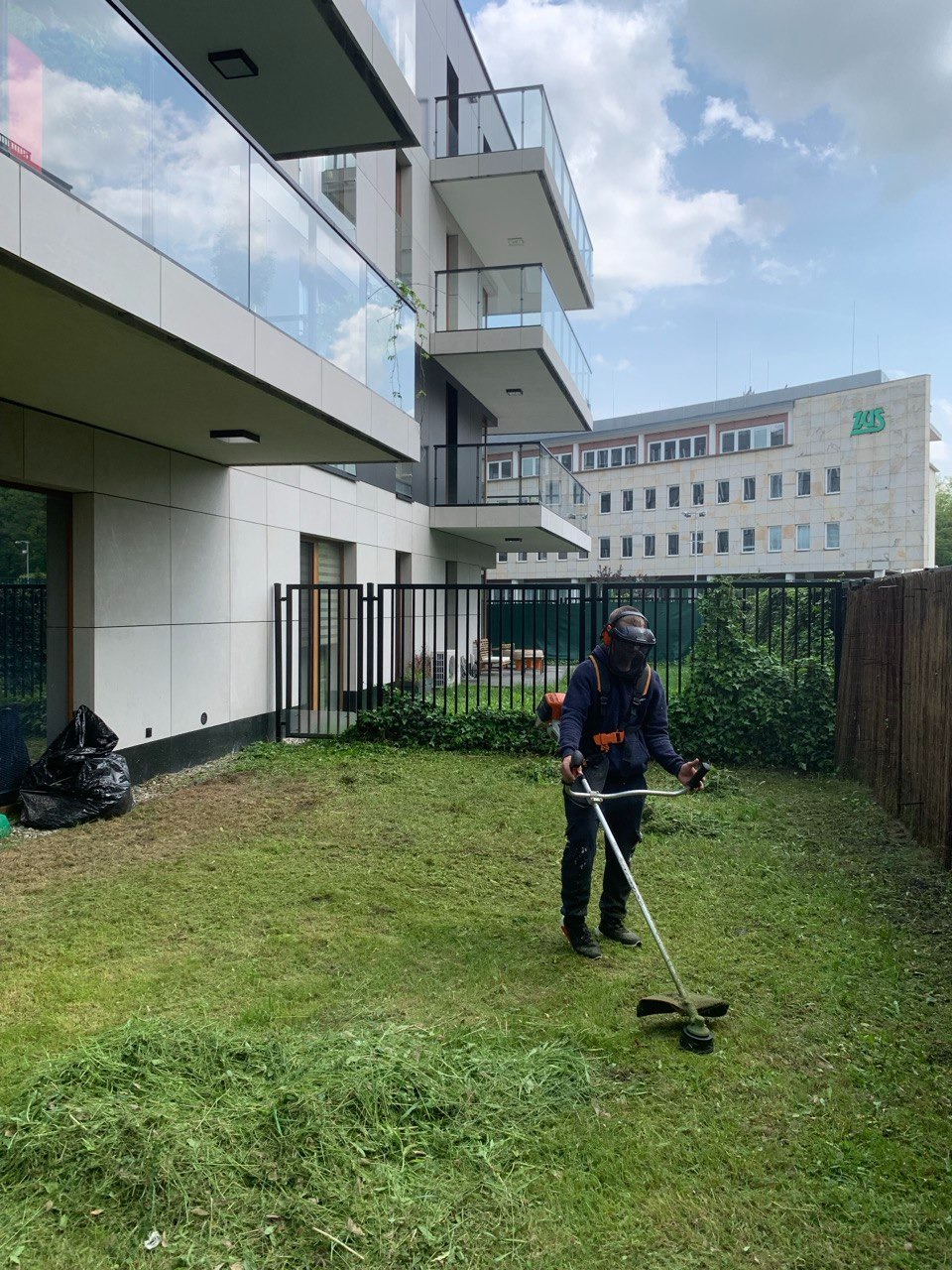 Pracownik w ochronnej masce i kombinezonie, używa podkaszarki spalinowej do wyrównywania trawy na tle nowoczesnego budynku z balkonami i budynkiem ZUS.