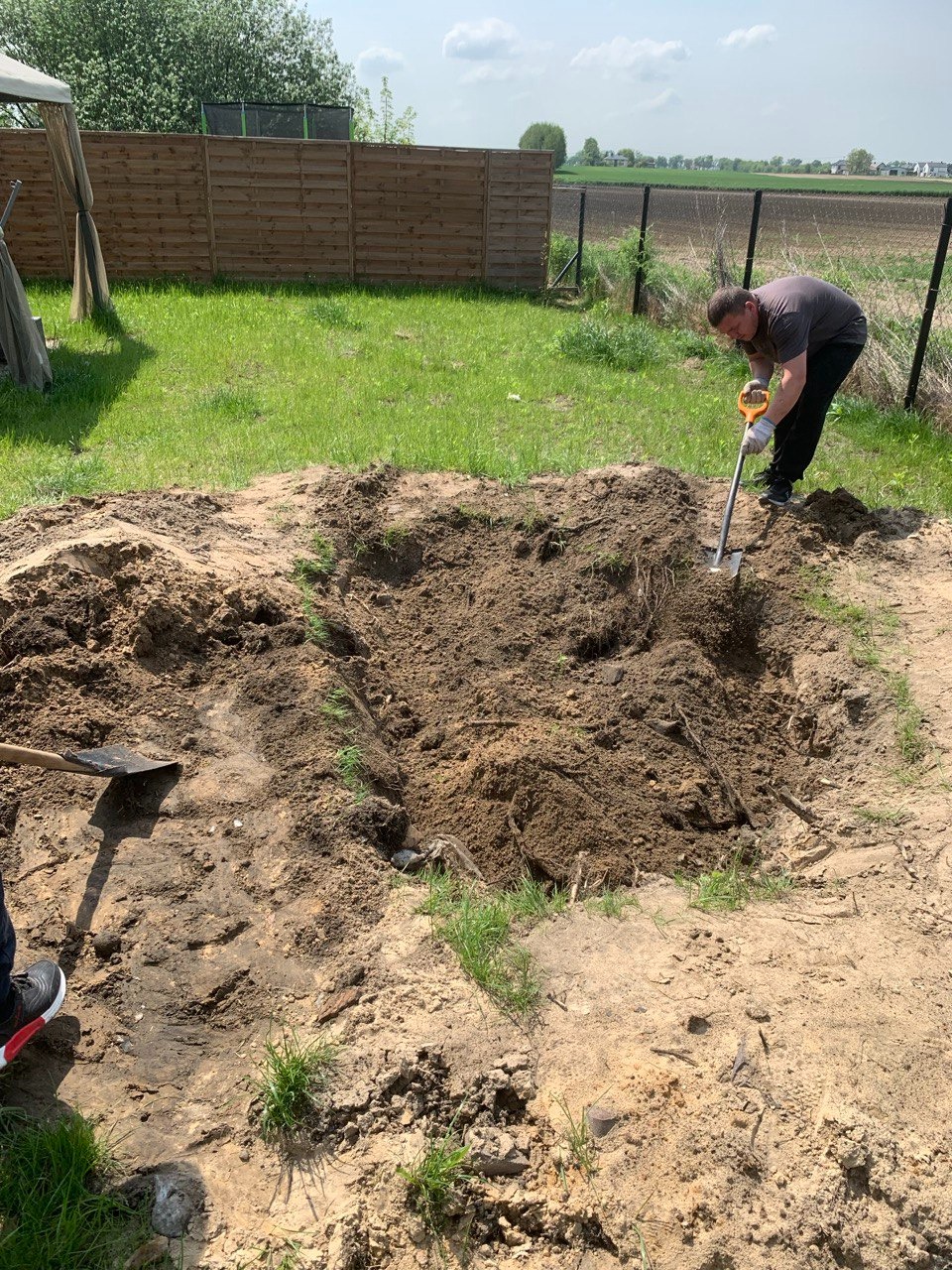 Pracownik kopie dół łopatą na terenie posesji z drewnianym ogrodzeniem w tle, przygotowując miejsce pod nasadzenia.