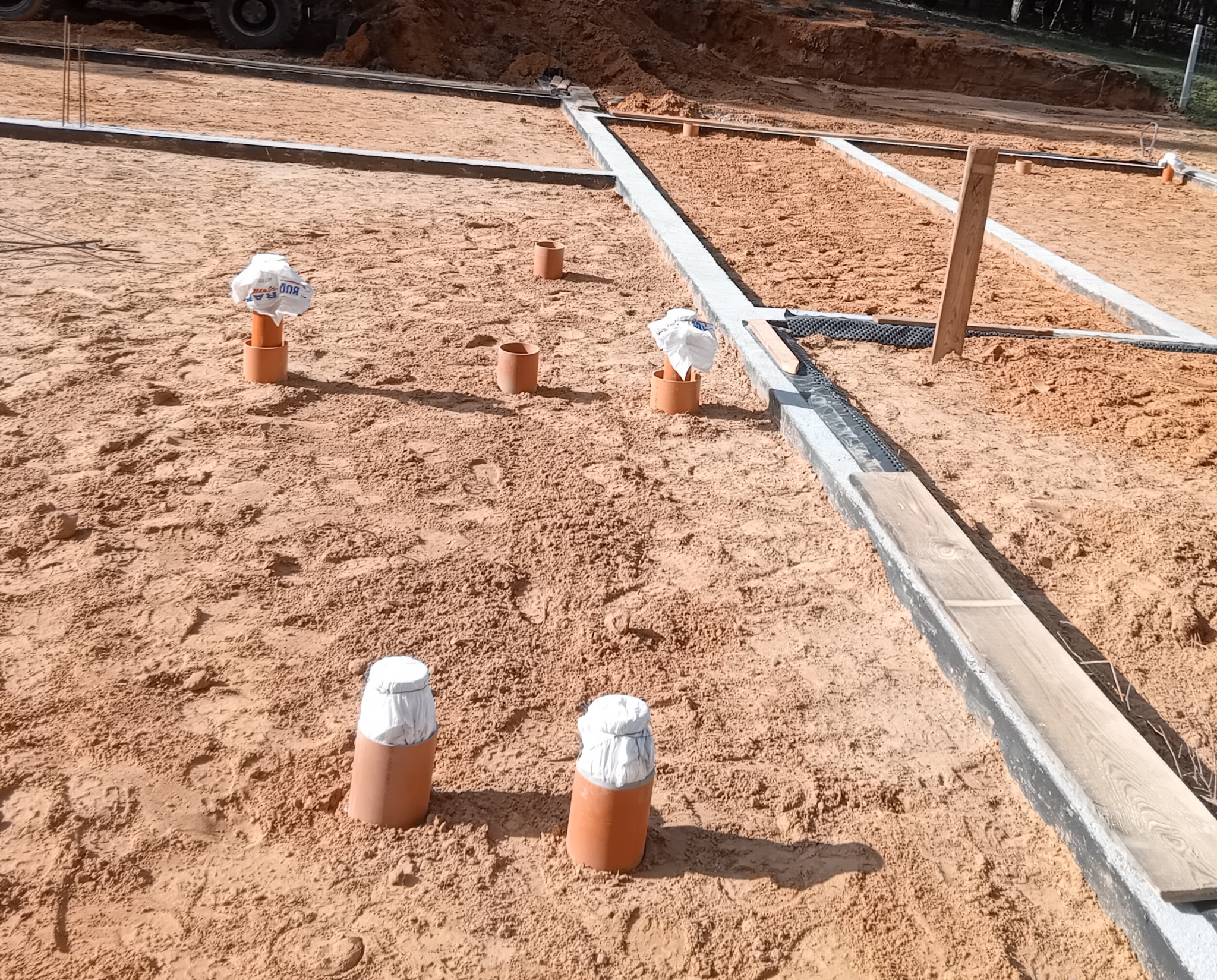 Przygotowanie terenu pod budowę z widocznymi rurami osłonowymi zabezpieczonymi folią, szalunkami i piaskiem.
