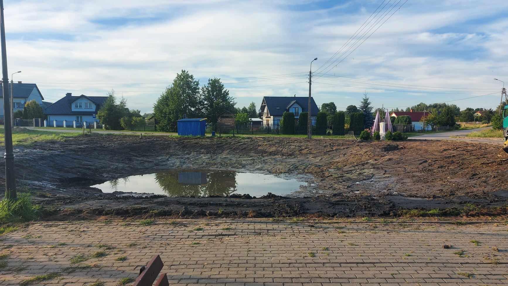 Wyrobisko ziemne wypełnione wodą, prawdopodobnie przygotowane pod oczko wodne, z widoczną kostką brukową na pierwszym planie i domami w tle.