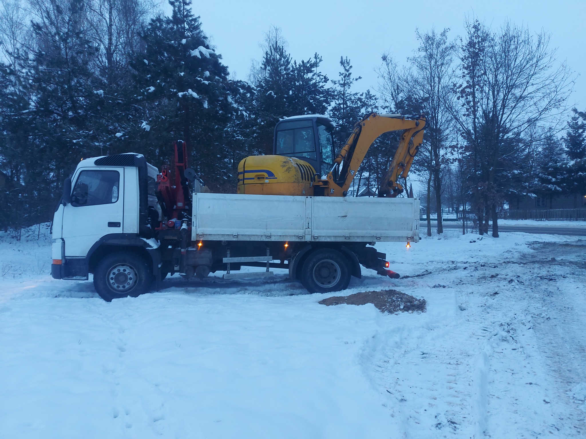 Biała ciężarówka z czerwoną hydrauliką przewożąca żółtą mini koparkę na tle zimowego krajobrazu z zasypanymi śniegiem drzewami.