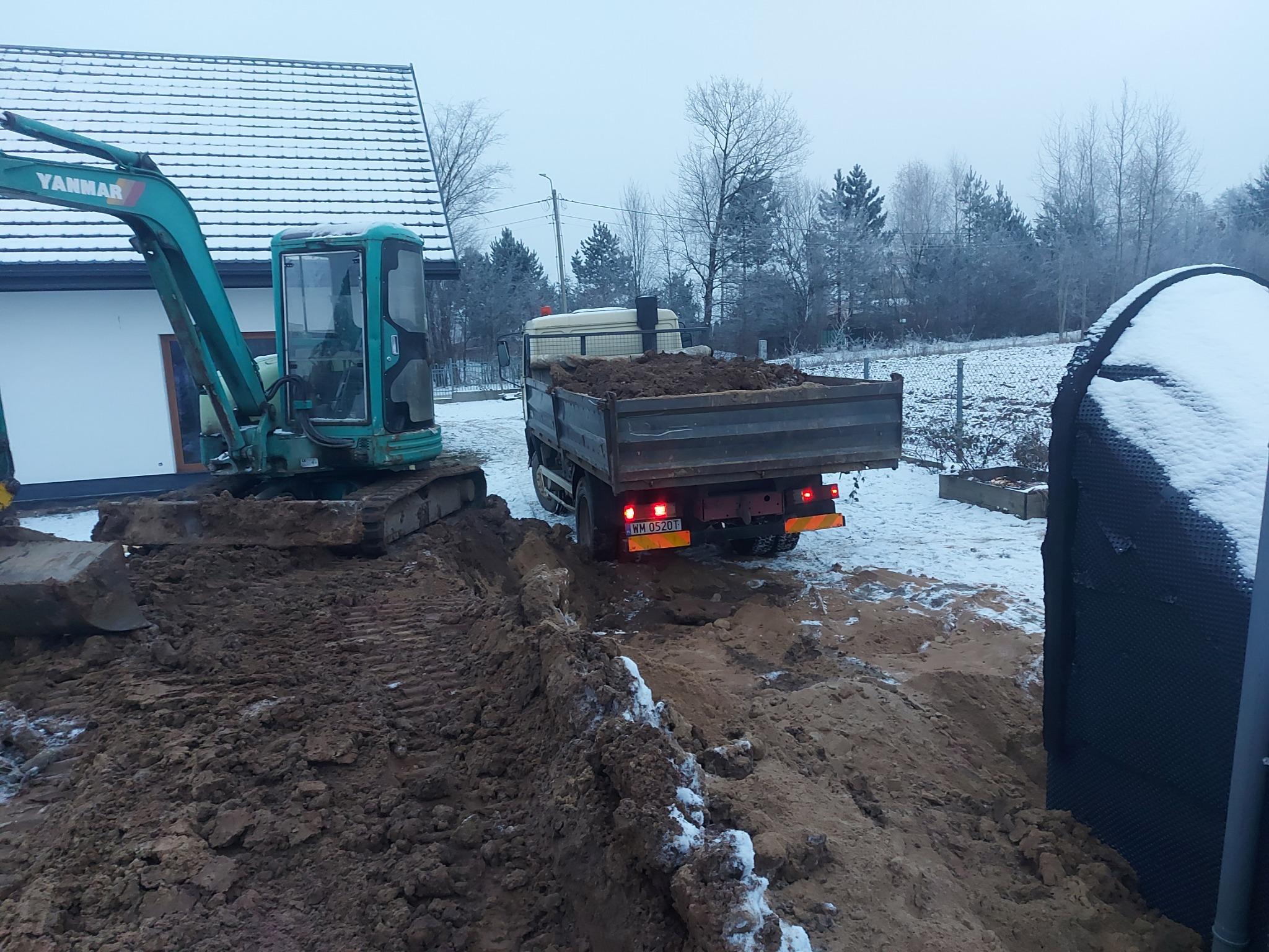 Nisko zawieszony, zielony minikoparka Yanmar obok wywrotki z ziemią, na placu budowy pokrytym śniegiem i błotem, w tle dom z ośnieżonym dachem.