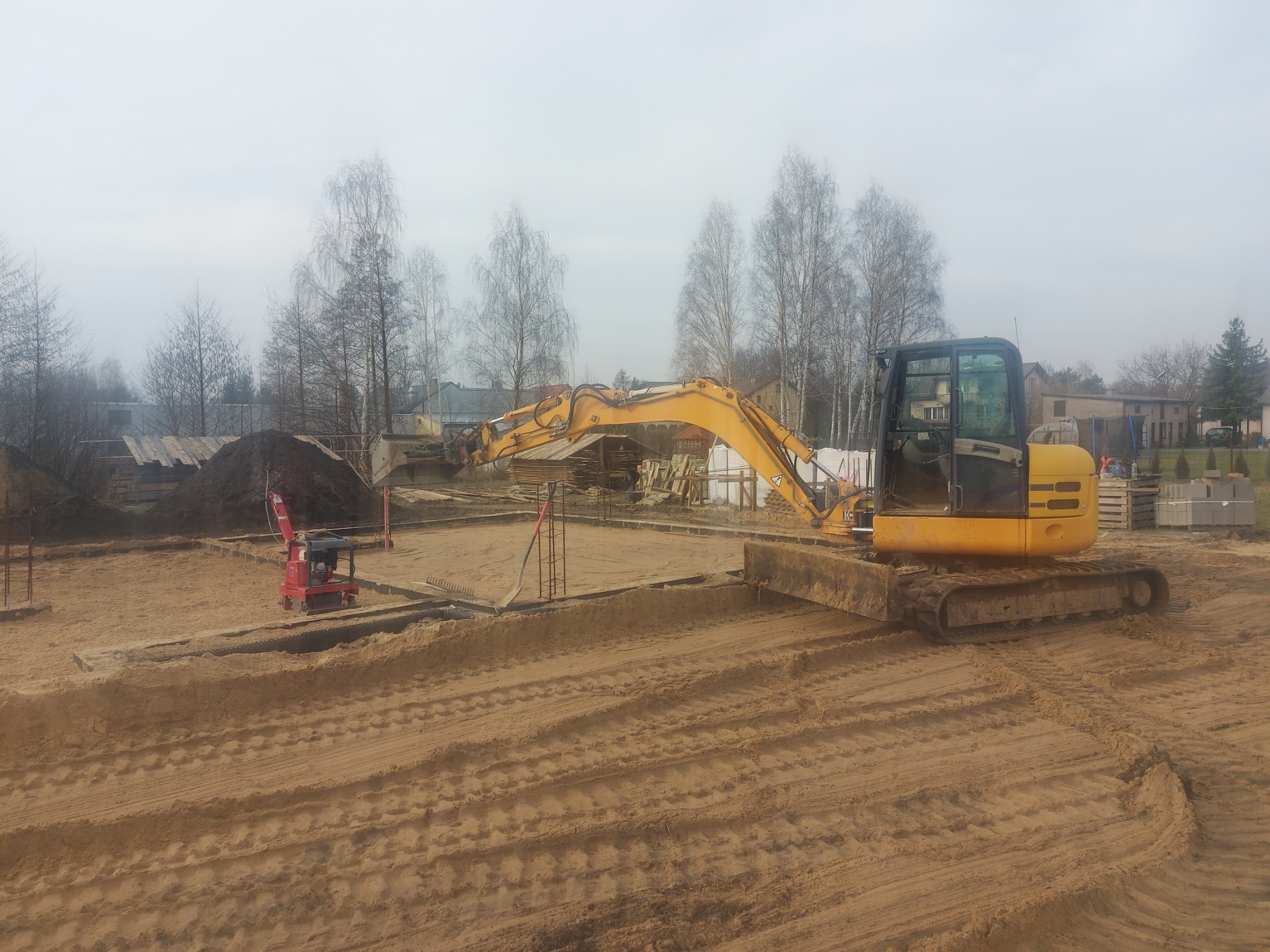 Żółta koparka wyrównuje teren pod fundamenty domu jednorodzinnego, widoczne zbrojenia i palety z bloczkami betonowymi w tle, pochmurny dzień.