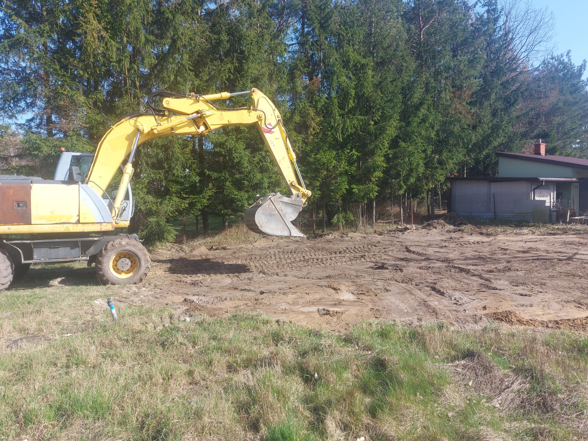 Żółta koparka kołowa na placu budowy, przygotowująca teren pod inwestycję, widoczne ślady opon na ubitej ziemi i trawa na pierwszym planie.