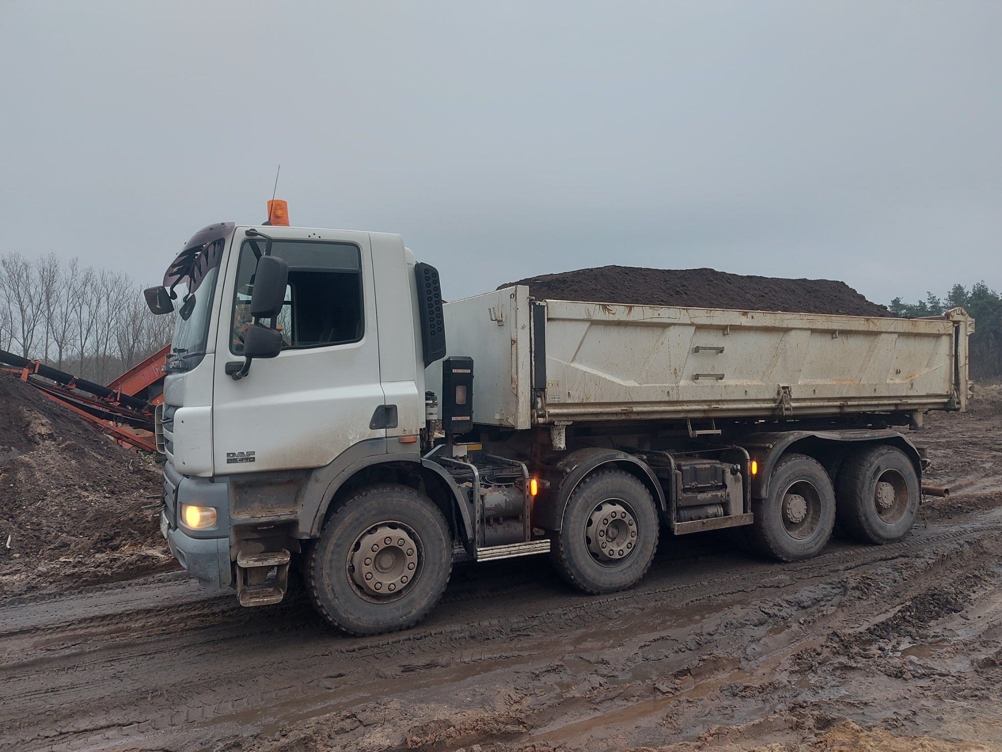 Ciężarówka DAF z przyczepą wypełnioną ziemią ogrodową na błotnistej drodze, w tle przesiewacz ziemi i drzewa.