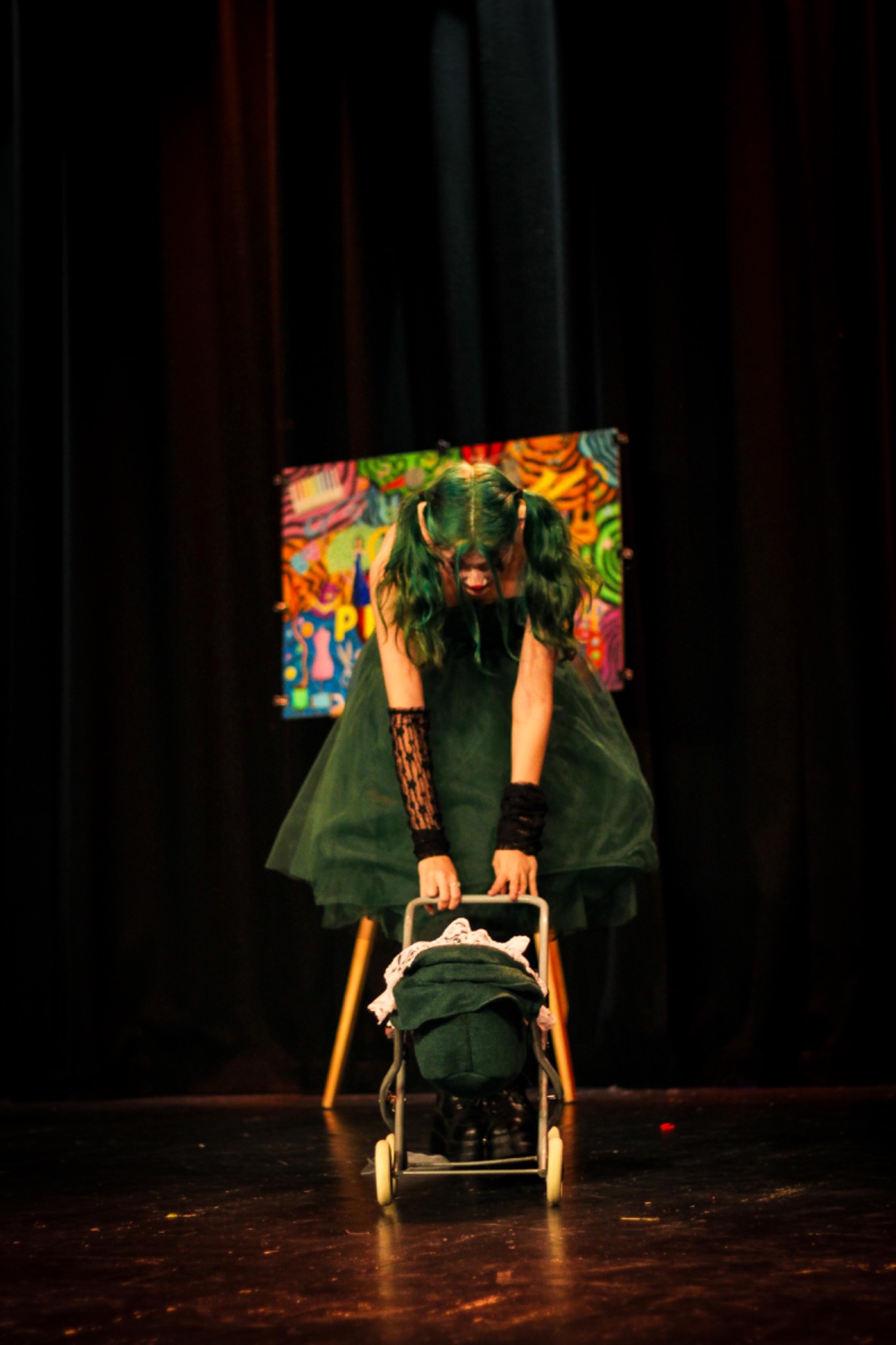 Kobieta w zielonej sukience i z zielonymi włosami stoi na scenie pochylona nad wózkiem dla lalek na tle kolorowej dekoracji.