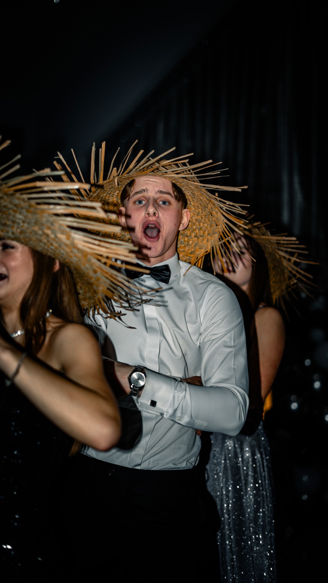 Młody mężczyzna w białej koszuli, czarnej muszce i słomkowym kapeluszu z szerokim rondem, z otwartymi ustami, w otoczeniu dwóch kobiet również w kapeluszach, na ciemnym tle podczas eventu.