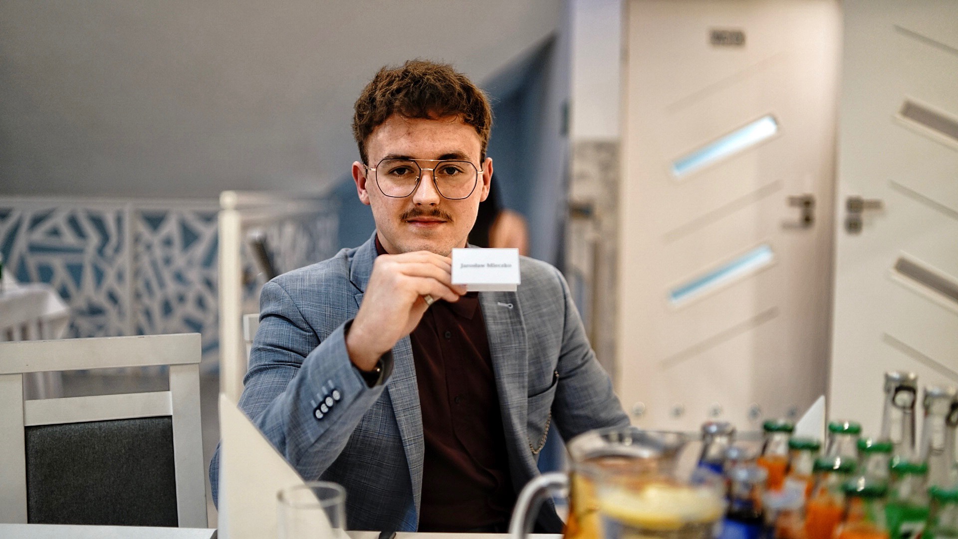 Mężczyzna w okularach i marynarce prezentuje wizytówkę z imieniem i nazwiskiem na tle sali bankietowej z napojami i dekoracjami.
