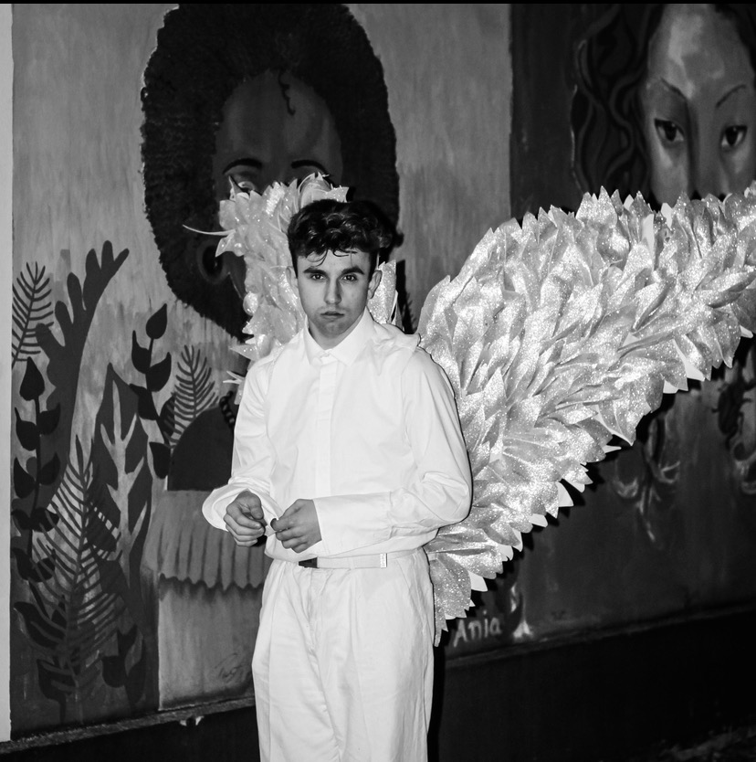 Młody mężczyzna w białym stroju i brokatowych skrzydłach anioła pozuje na tle malowidła ściennego z portretami i roślinami.