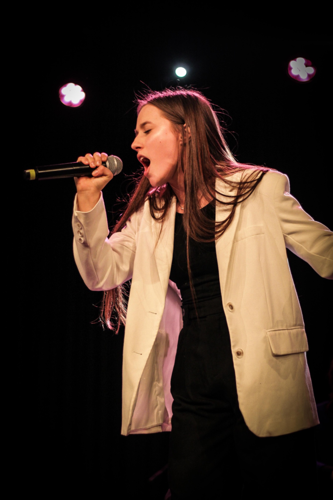Młoda kobieta w białym żakiecie śpiewa do mikrofonu na scenie z ciemnym tłem i dekoracyjnymi światłami.
