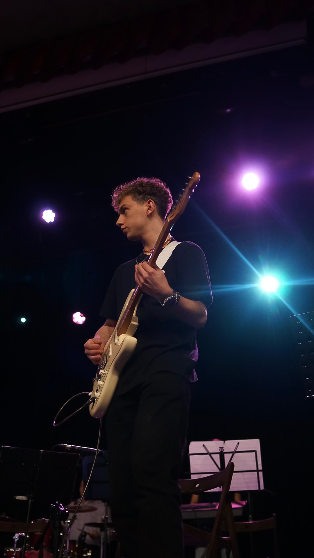 Młody muzyk z kręconymi włosami gra na białej gitarze elektrycznej na scenie oświetlonej fioletowymi i zielonymi reflektorami, w tle widoczne nuty, perkusja i klawisze.