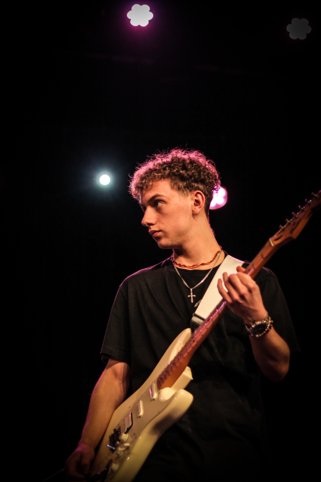 Młody gitarzysta z kręconymi włosami, w czarnym T-shircie, grający na białej gitarze elektrycznej podczas koncertu, oświetlony reflektorami.