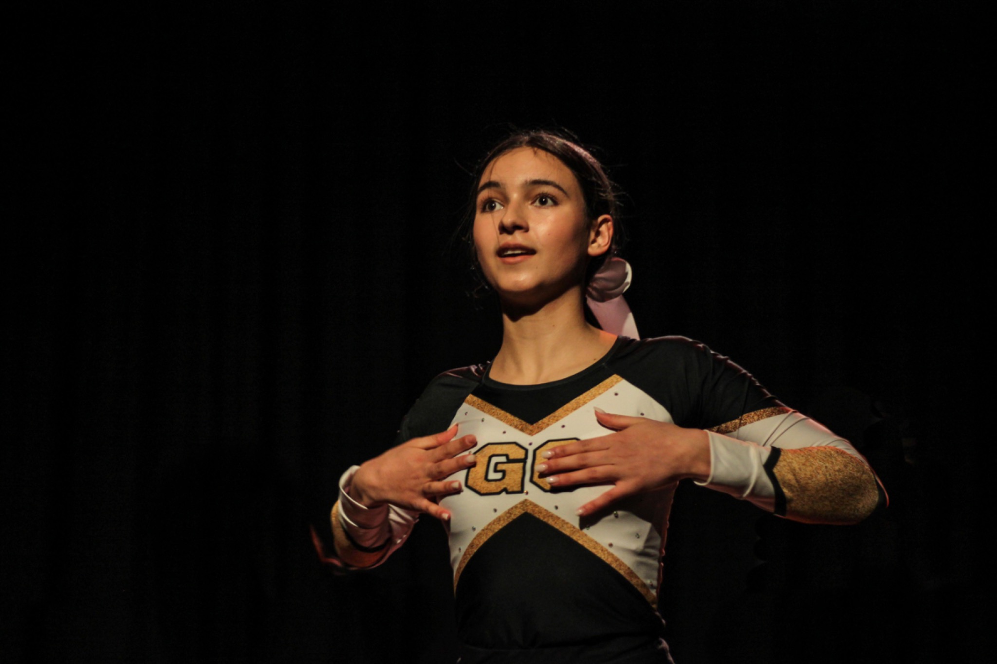 Młoda cheerleaderka w stroju z literami 'GC' na scenie, z rękami ułożonymi na piersi, patrzy w górę, oświetlona punktowym światłem na ciemnym tle.
