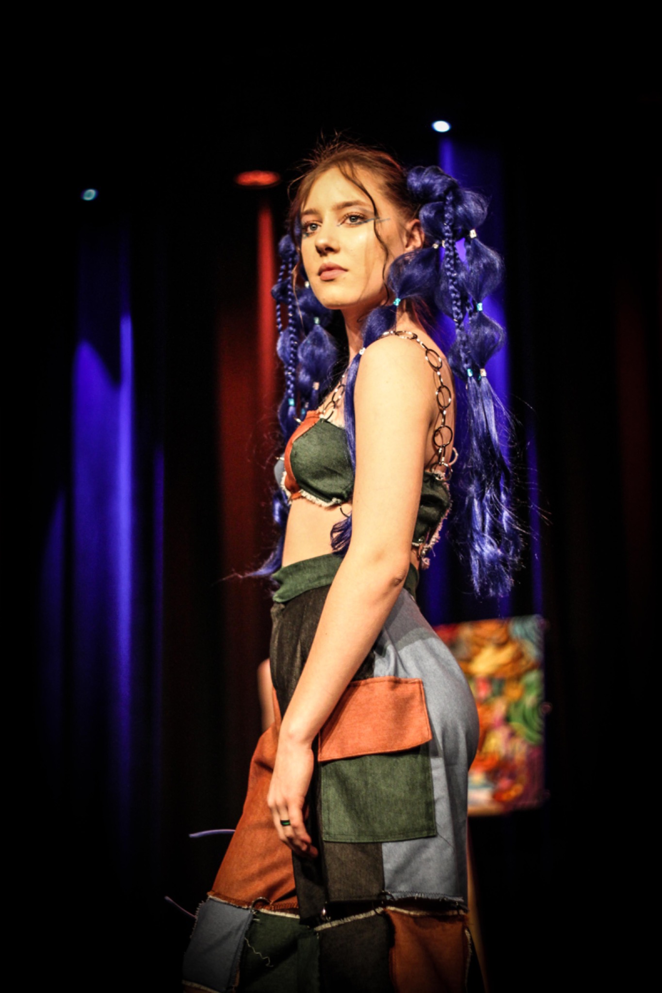 Młoda modelka z niebieskimi warkoczykami, w patchworkowym stroju, pozuje na tle ciemnej sceny z kolorowymi światłami.