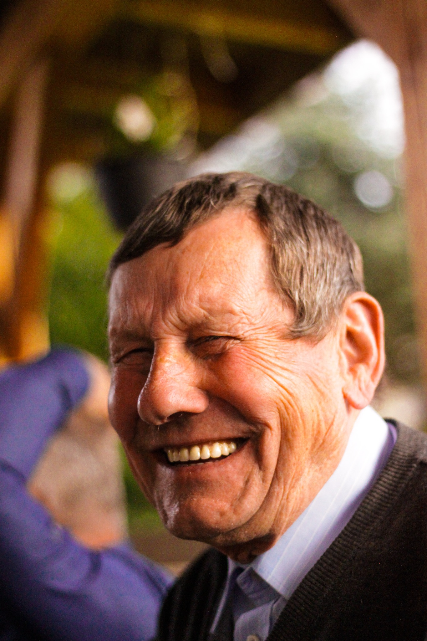 Portret uśmiechniętego starszego mężczyzny z przymkniętymi oczami, ukazujący zmarszczki i promienny uśmiech, ubrany w koszulę i sweter w naturalnym oświetleniu.