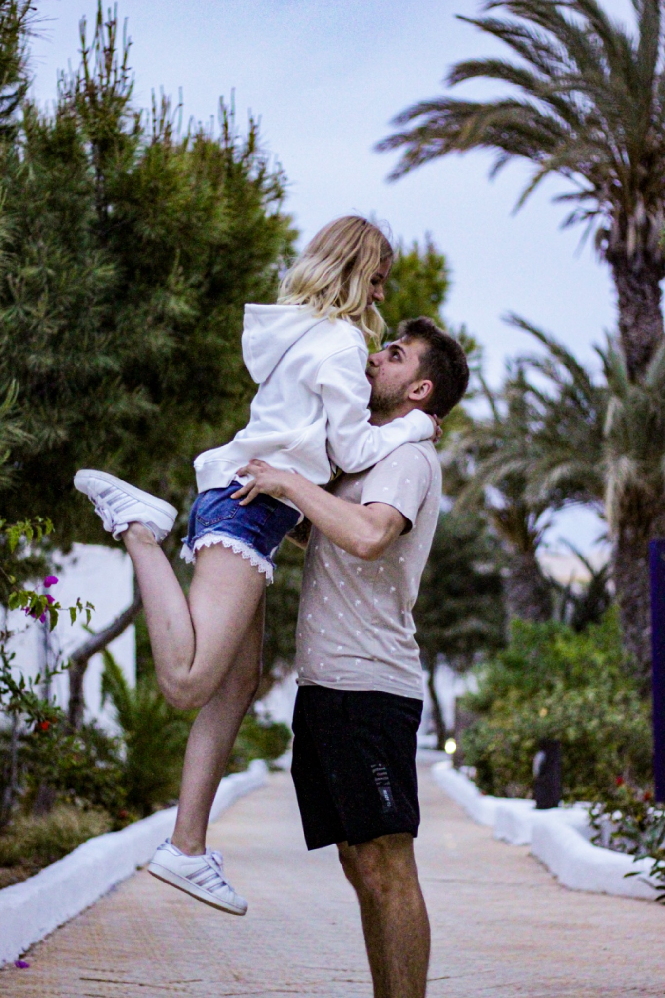 Mężczyzna podnosi blondynkę w białej bluzie i dżinsowych szortach na tle tropikalnej roślinności.