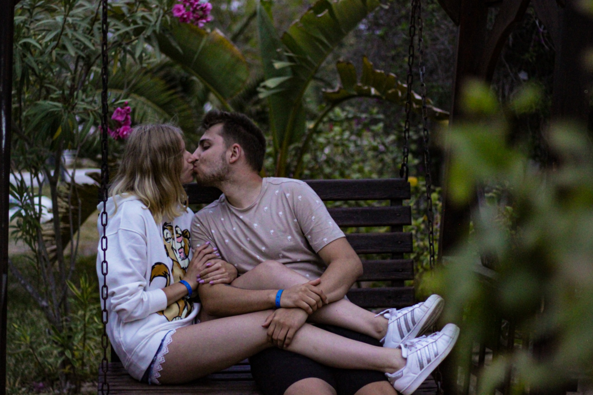Para całująca się na drewnianej huśtawce w otoczeniu tropikalnej roślinności i różowych kwiatów.