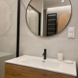 Nowoczesna łazienka z umywalką nablatową na drewnianej szafce, czarną armaturą i okrągłym lustrem, widoczna kabina prysznicowa z czarnymi profilami i grzejnik drabinkowy w odbiciu lustra.