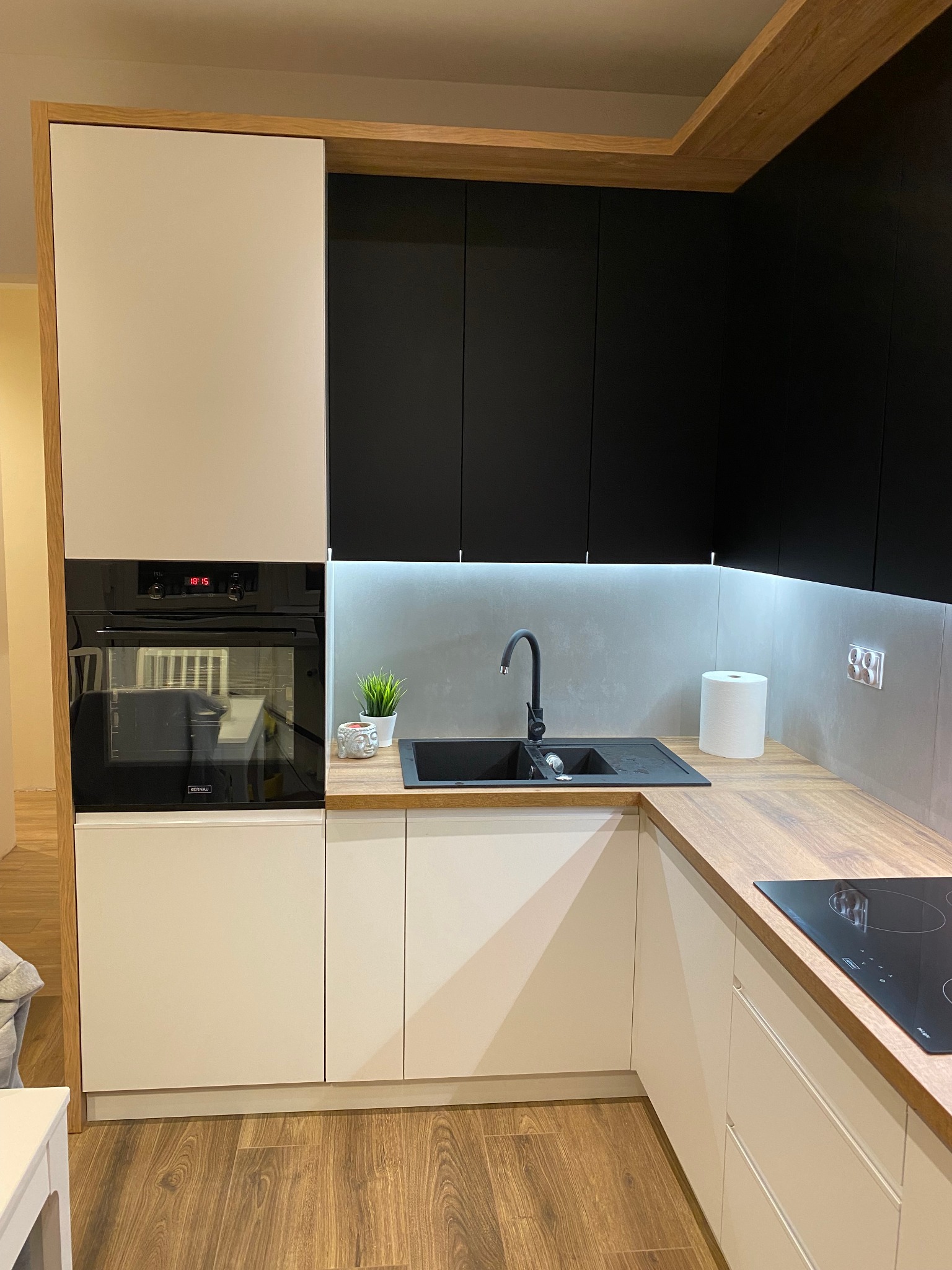 Nowoczesna kuchnia z białymi i czarnymi szafkami, drewnianym blatem i czarnym zlewem, oświetlona listwą LED pod szafkami wiszącymi.