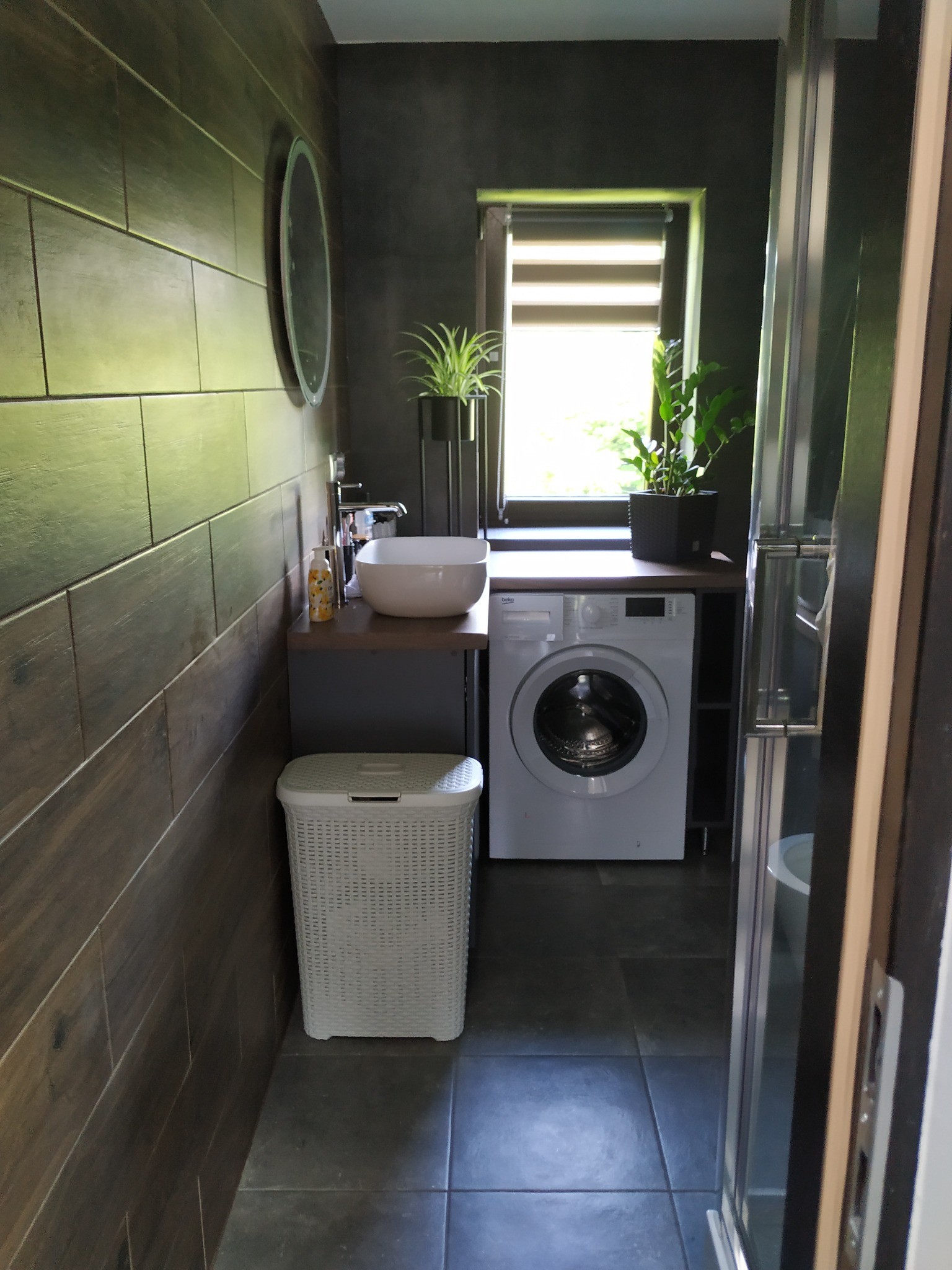 Nowoczesna łazienka z drewnopodobnymi płytkami na ścianie, owalną umywalką nablatową, pralką pod blatem i roślinami w doniczkach na parapecie.