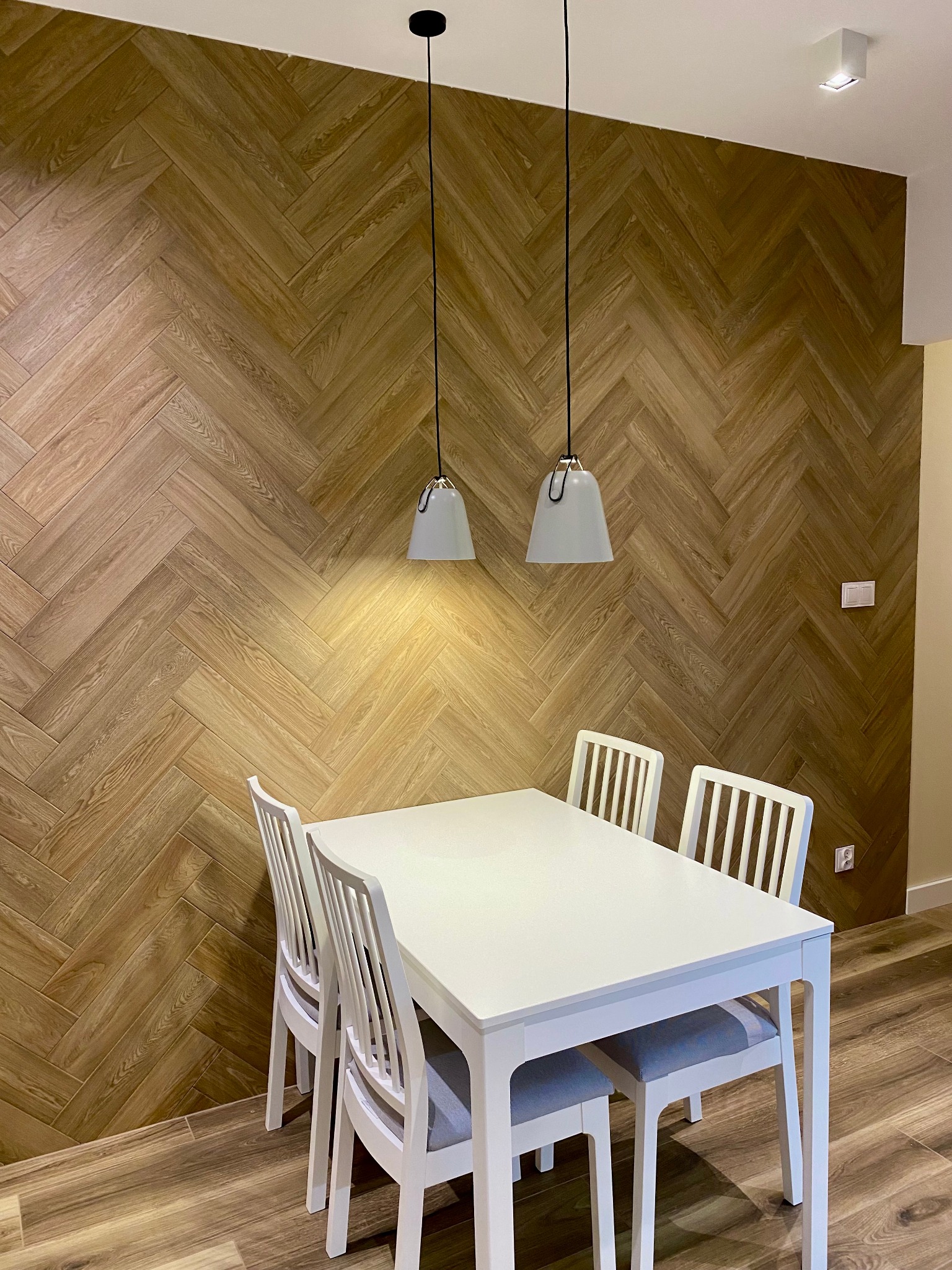Jadalnia z drewnianą ścianą ułożoną w jodełkę, oświetlona dwoma wiszącymi lampami z białymi kloszami, biały stół i krzesła, podłoga imitująca drewno.