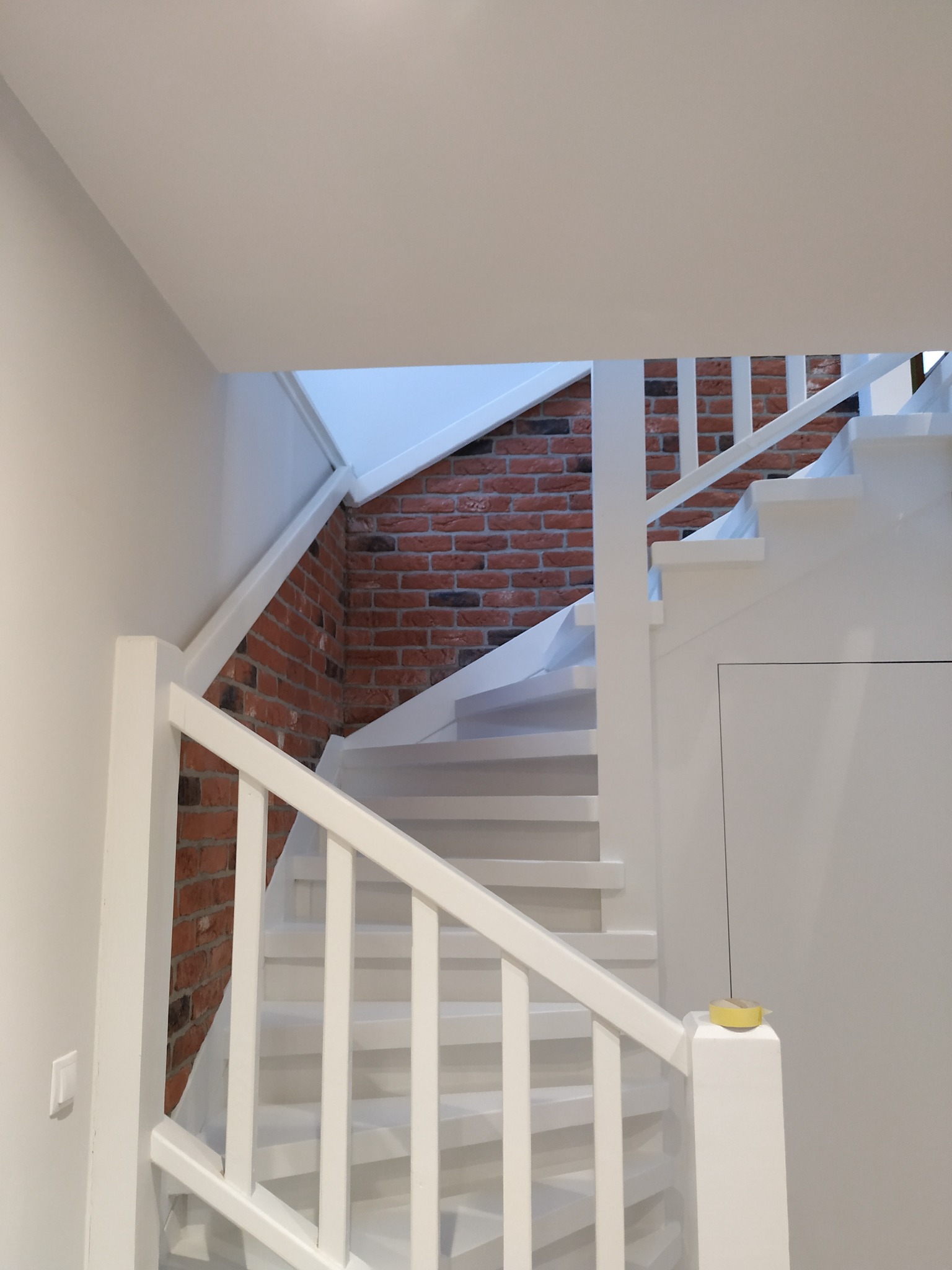 Białe schody z drewnianą balustradą i ceglaną ścianą w tle, zabezpieczone żółtą taśmą malarską na słupku.
