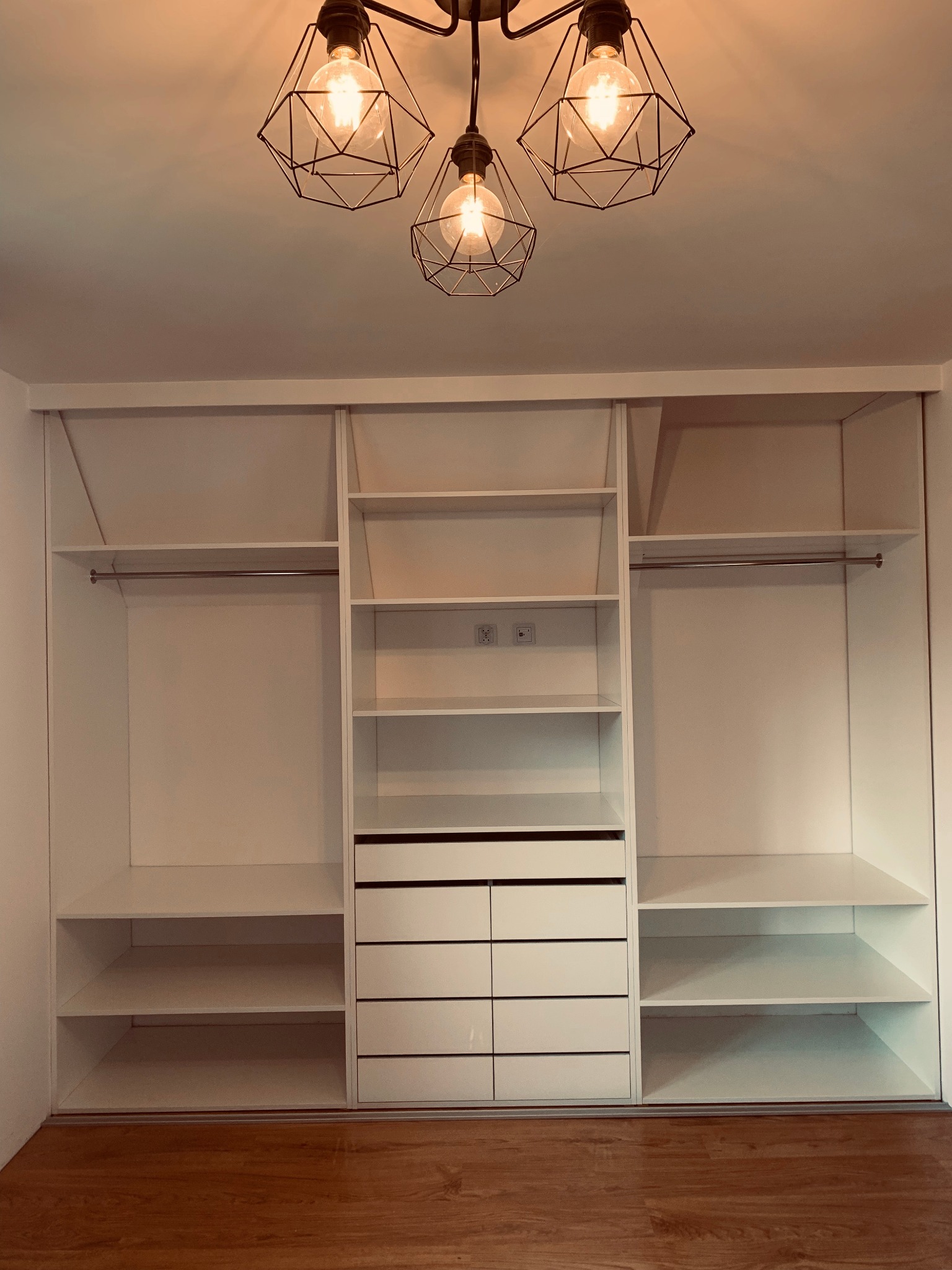 Biała, pojemna szafa wnękowa z przesuwnymi drzwiami i szufladami, oświetlona designerską lampą z trzema żarówkami na tle parkietu.