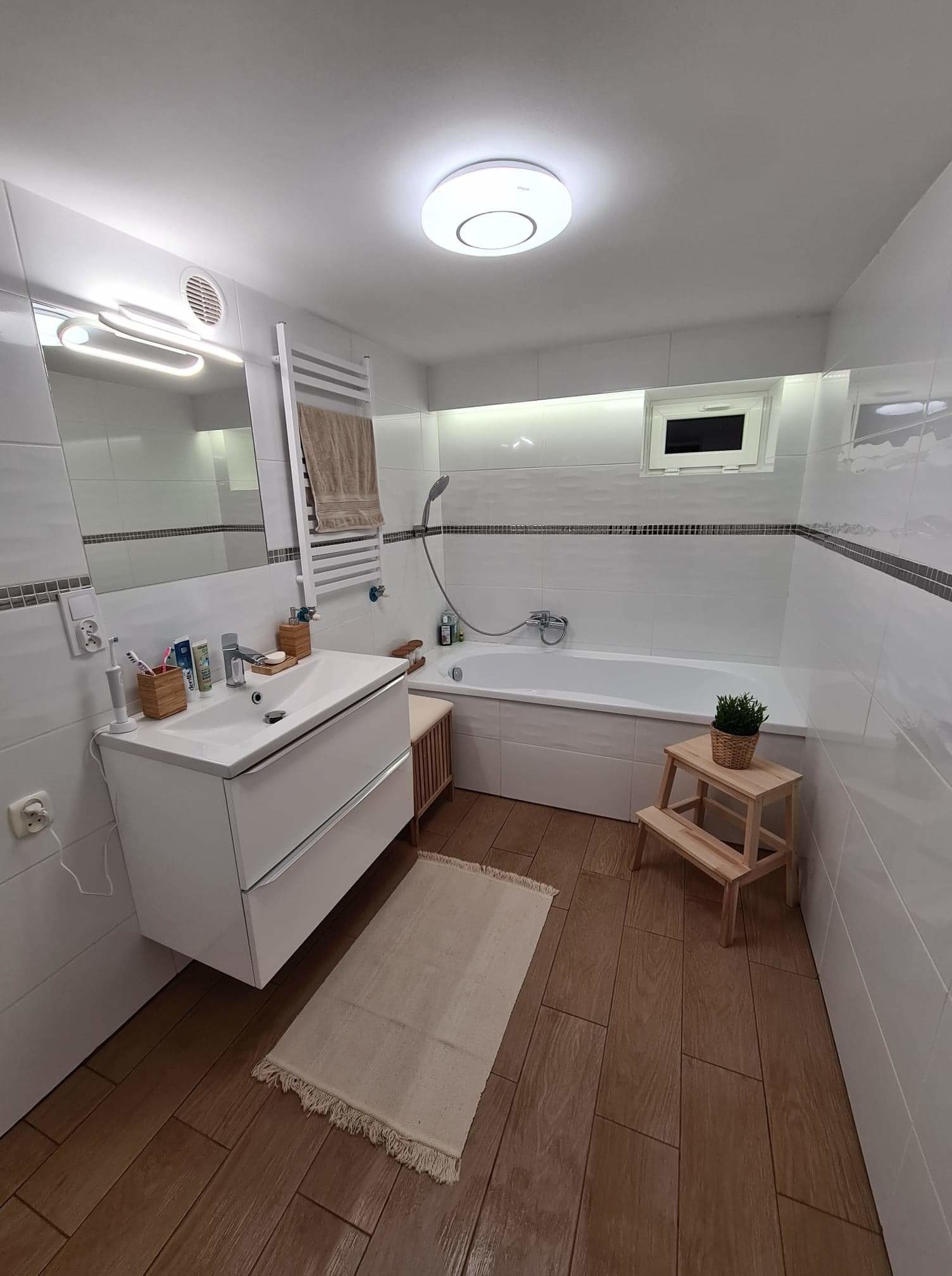 Jasna łazienka po remoncie z białymi płytkami, drewnianą podłogą, umywalką z szafką, wanną, drabinkowym grzejnikiem i drewnianym stołkiem z rośliną.