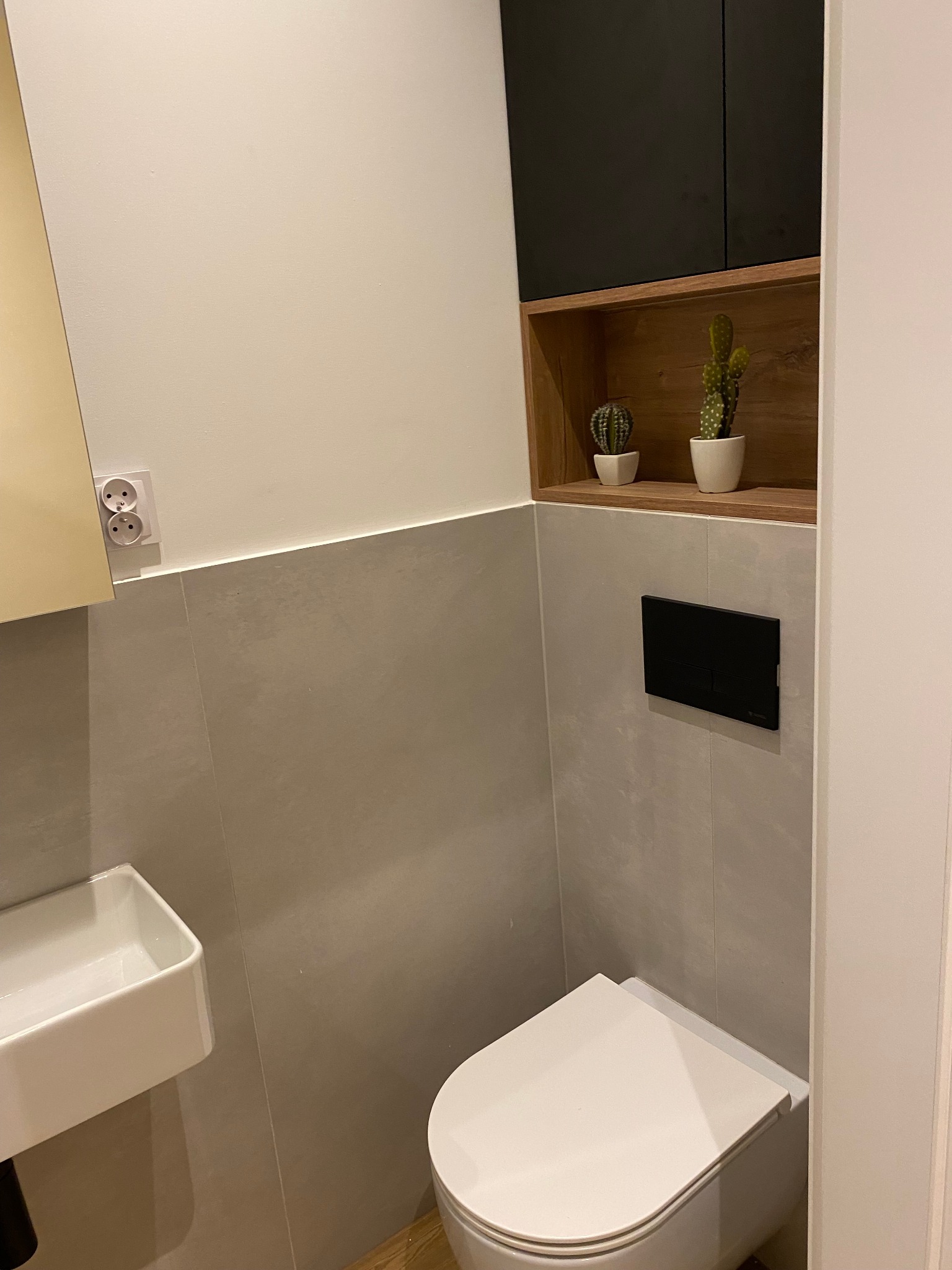 Nowoczesna toaleta z szarymi płytkami, wiszącą miską WC, umywalką i półką z dwoma kaktusami w białych doniczkach.