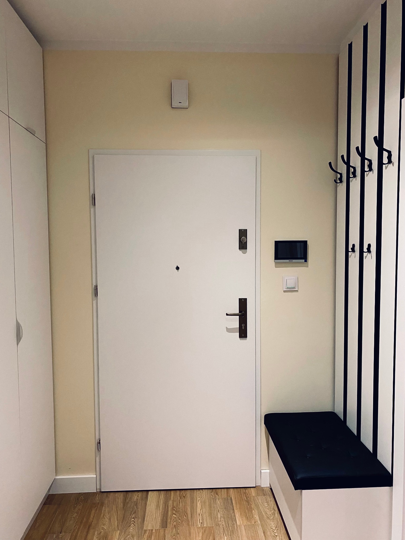 Przedpokój z białą szafą, drzwiami wejściowymi, panelem z wieszakami w czarno-białe pasy oraz czarną pufą na białej skrzyni.