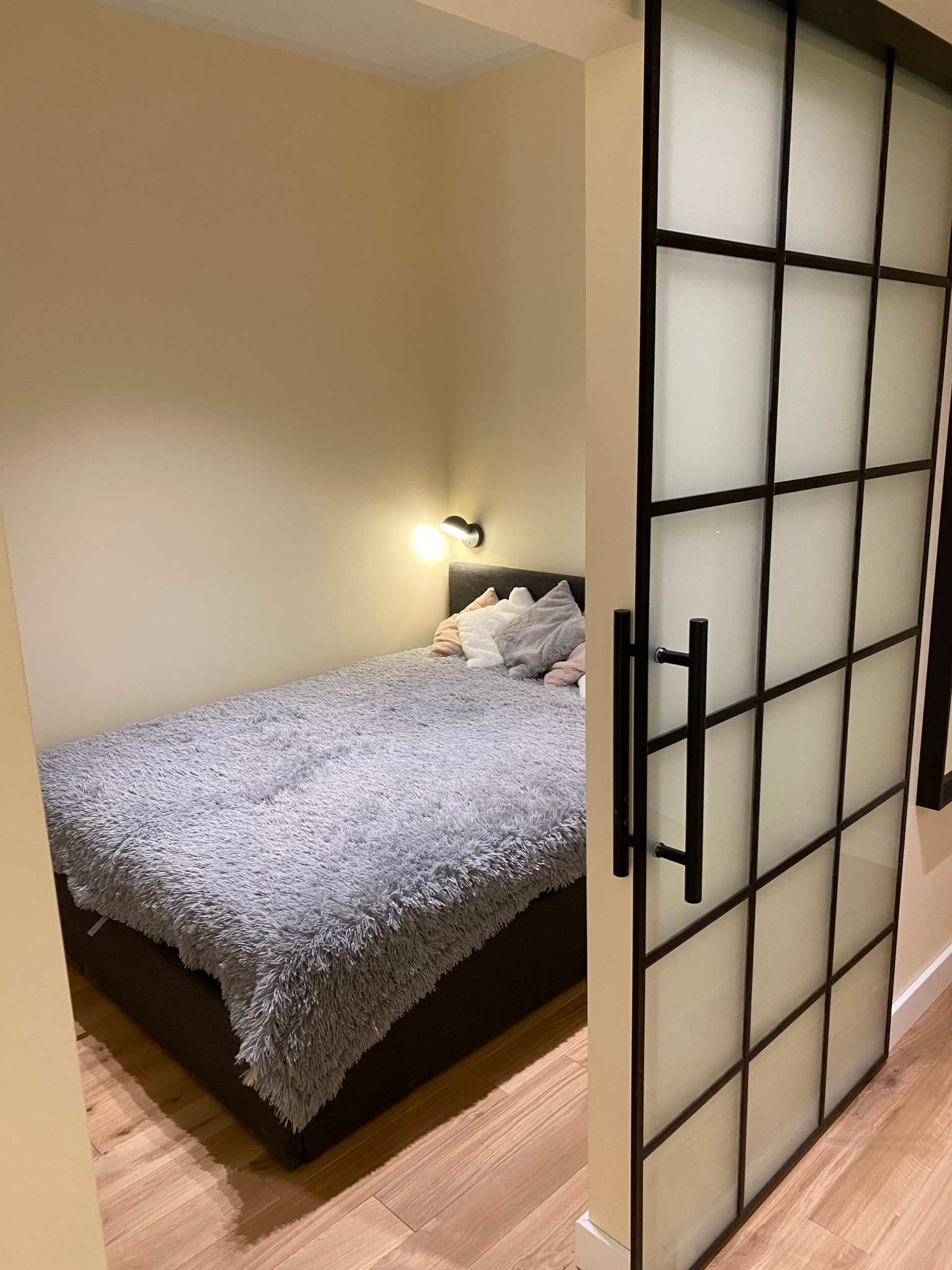 Niewielka sypialnia z łóżkiem przy ścianie, oświetlona kinkietem, z przesuwnymi drzwiami w stylu industrialnym z matowego szkła i czarnych szprosów.