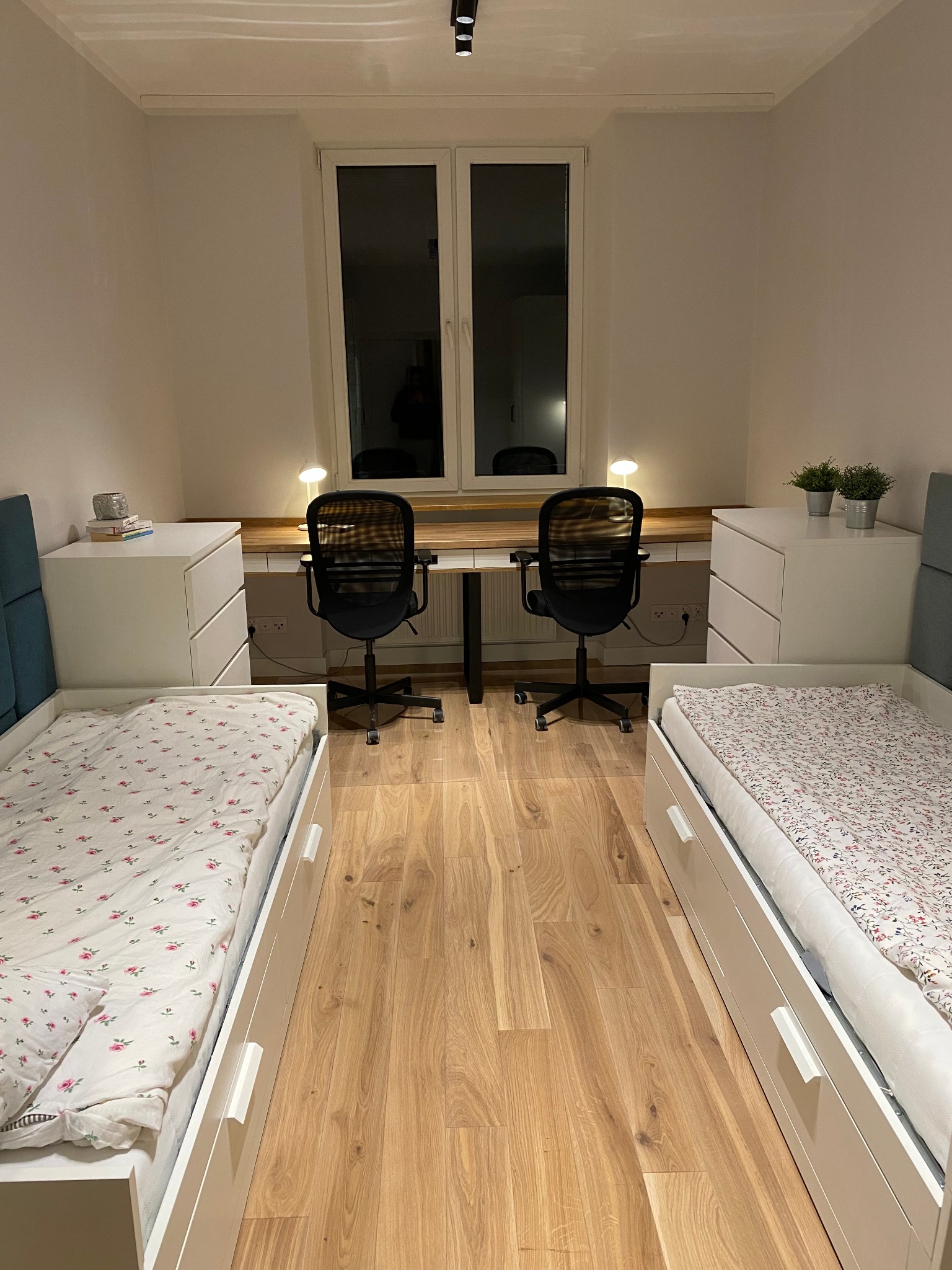 Pokój z dwoma łóżkami z szufladami i biurkami pod oknem, drewniana podłoga, białe ściany, minimalistyczny styl.