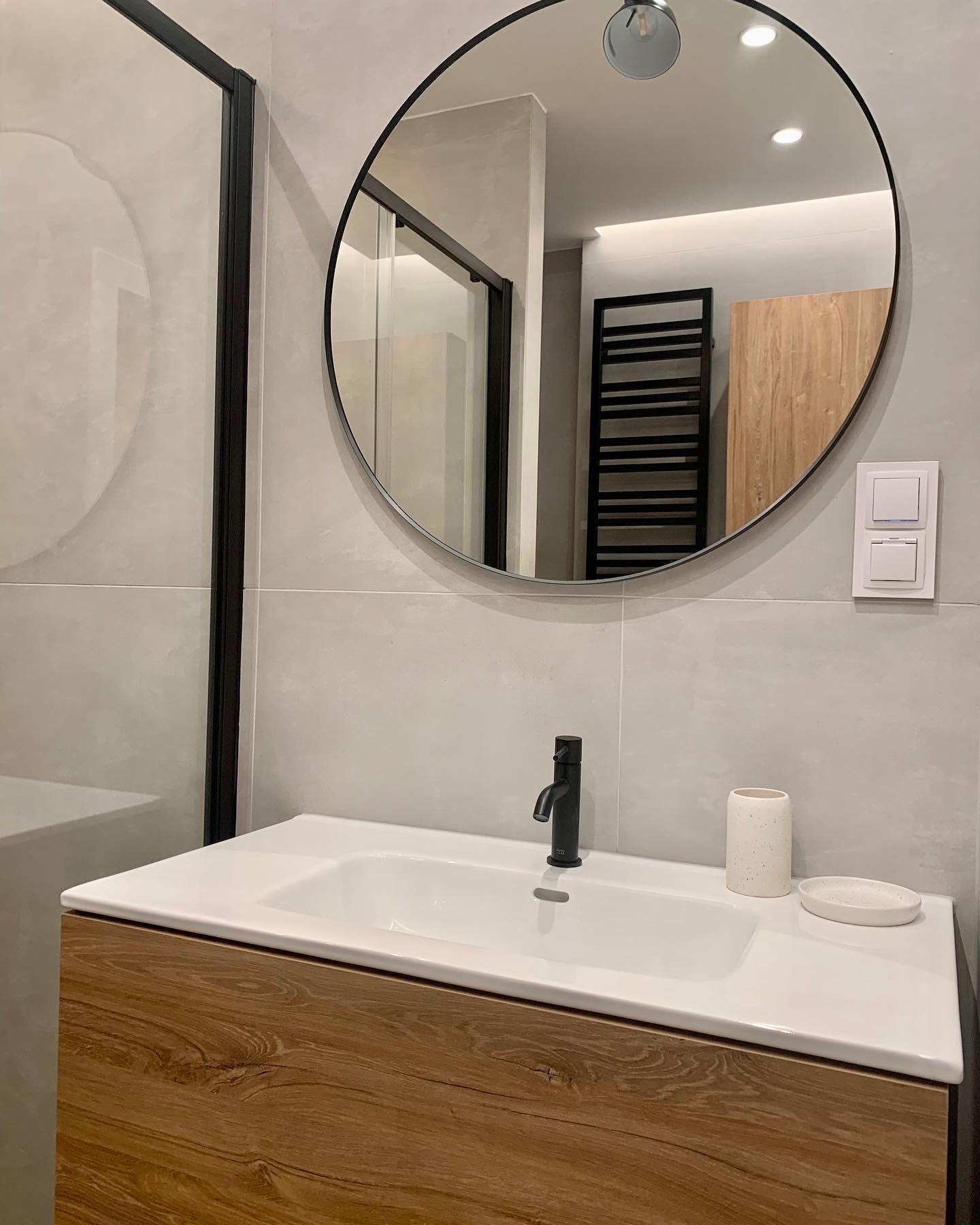 Nowoczesna łazienka z umywalką nablatową na drewnianej szafce, czarną armaturą i okrągłym lustrem, widoczna kabina prysznicowa z czarnymi profilami i grzejnik drabinkowy w odbiciu lustra.