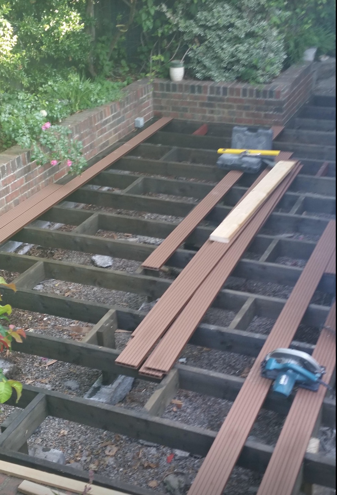 Konstrukcja drewnianego tarasu w ogrodzie, z widocznymi deskami kompozytowymi i narzędziami, etap budowy przy ceglanym murku z roślinami.