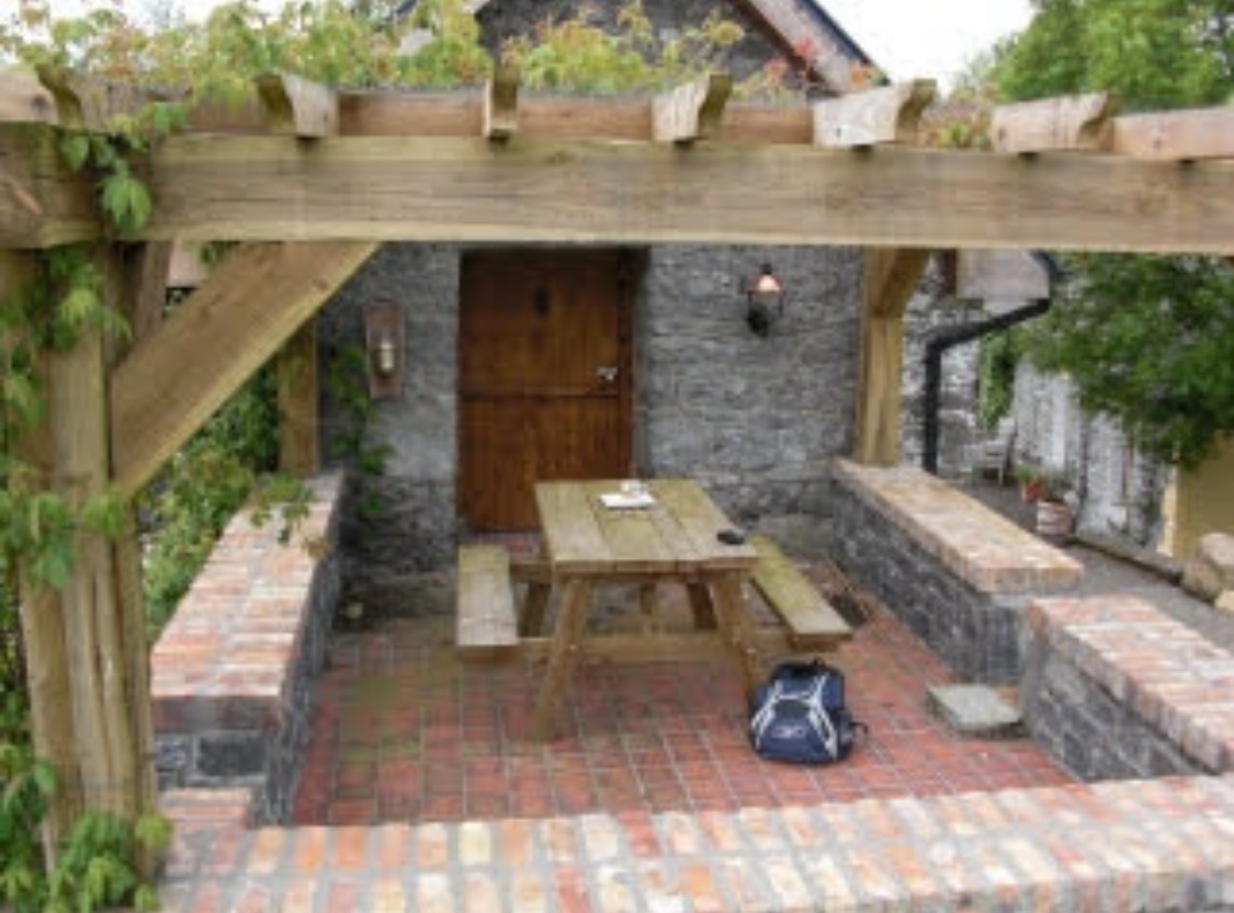 Zadaszony taras z ceglaną podłogą i kamiennymi ścianami. Drewniany stół piknikowy, plecak i lampa ścienna dodają uroku.