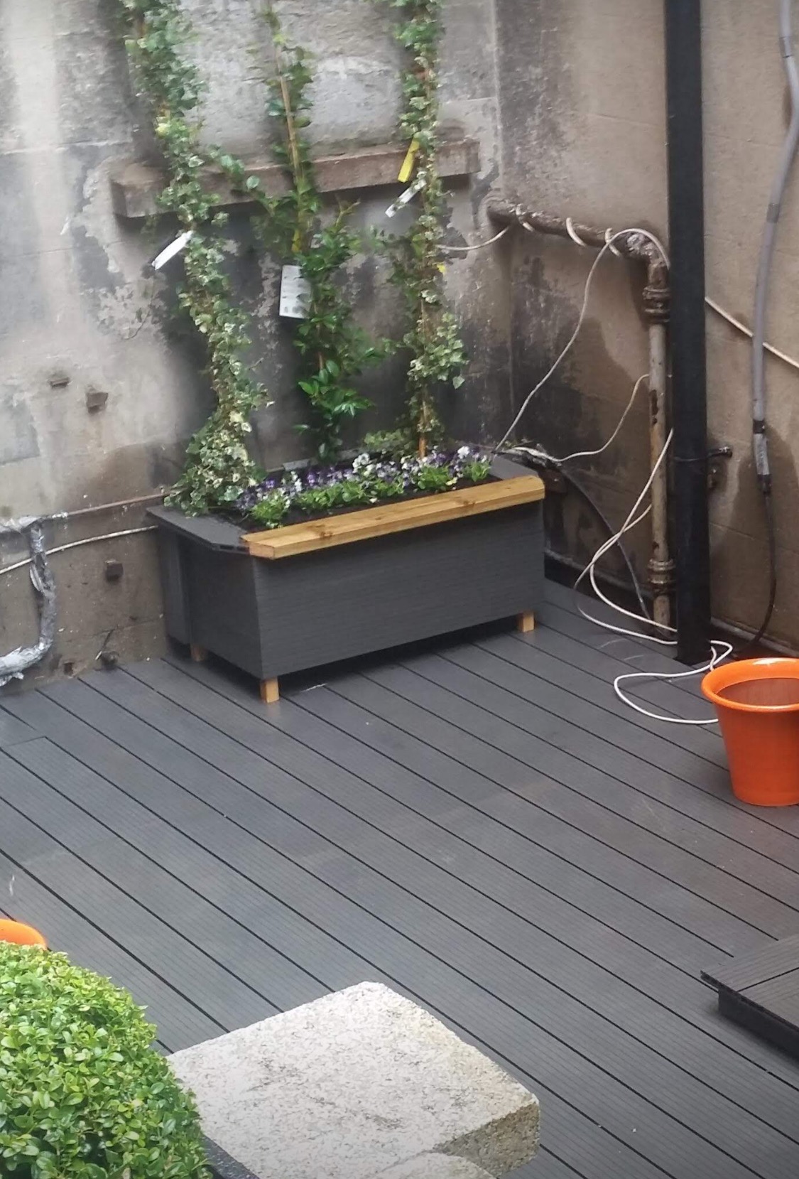 Szara donica z kwiatami na drewnianym tarasie balkonowym, z bluszczem pnącym się po ścianie. Elementy wykończenia balkonu w szarościach i naturalnym drewnie.