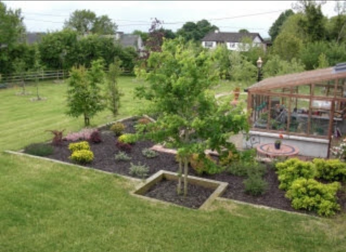 Uporządkowany ogród z zielonym trawnikiem, rabatami kwiatowymi i małą szklarnią, tworzący harmonijną przestrzeń.