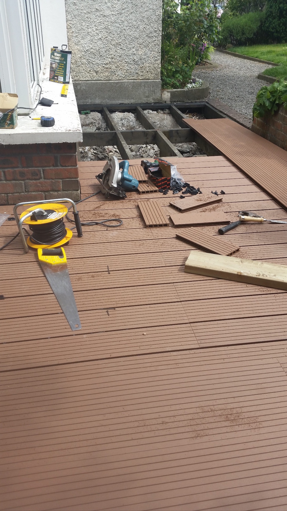 Montaż brązowego tarasu kompozytowego na zewnątrz budynku. Widoczne narzędzia: piła, przedłużacz, młotek. Częściowo ułożone deski i konstrukcja nośna.