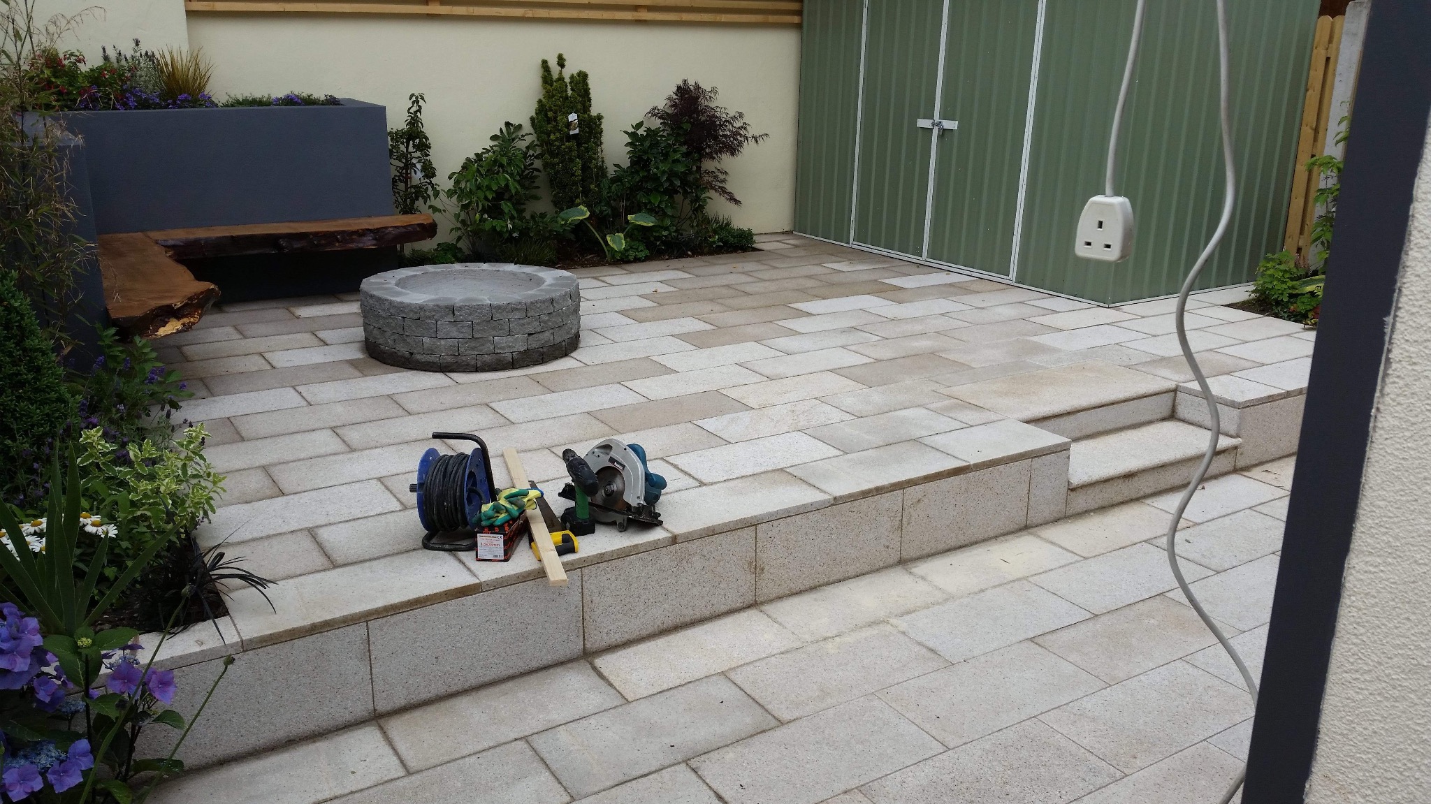 Ukończony taras z jasnej kostki brukowej z elementami małej architektury i narzędziami pracy, widok z góry, kabel zwisa z kontaktu.