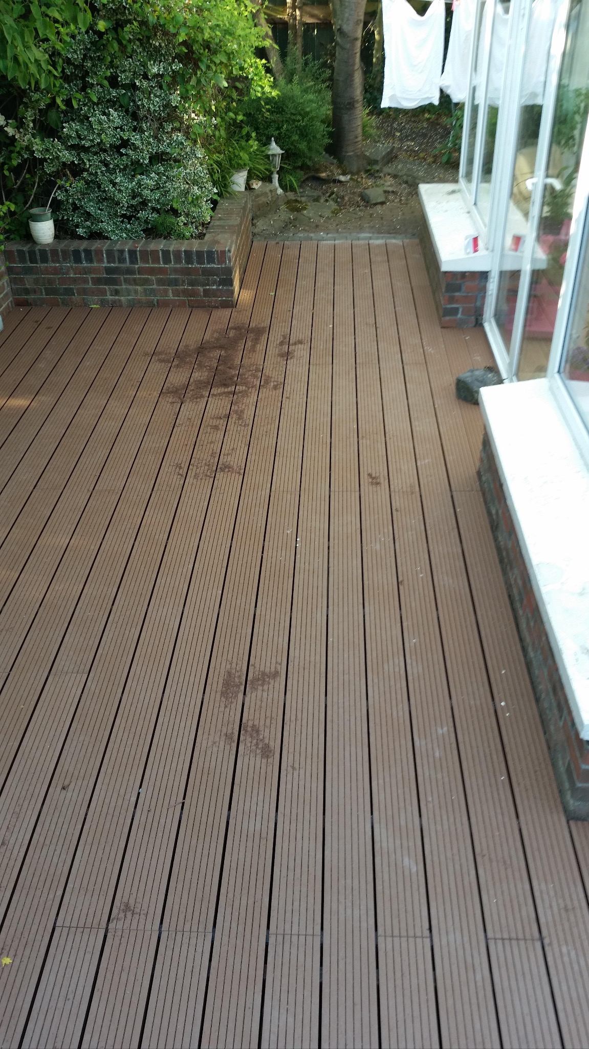 Nowy taras z desek kompozytowych w ogrodzie, widoczne ślady zabrudzeń. Elementy zieleni w tle, białe pranie suszy się na sznurku.